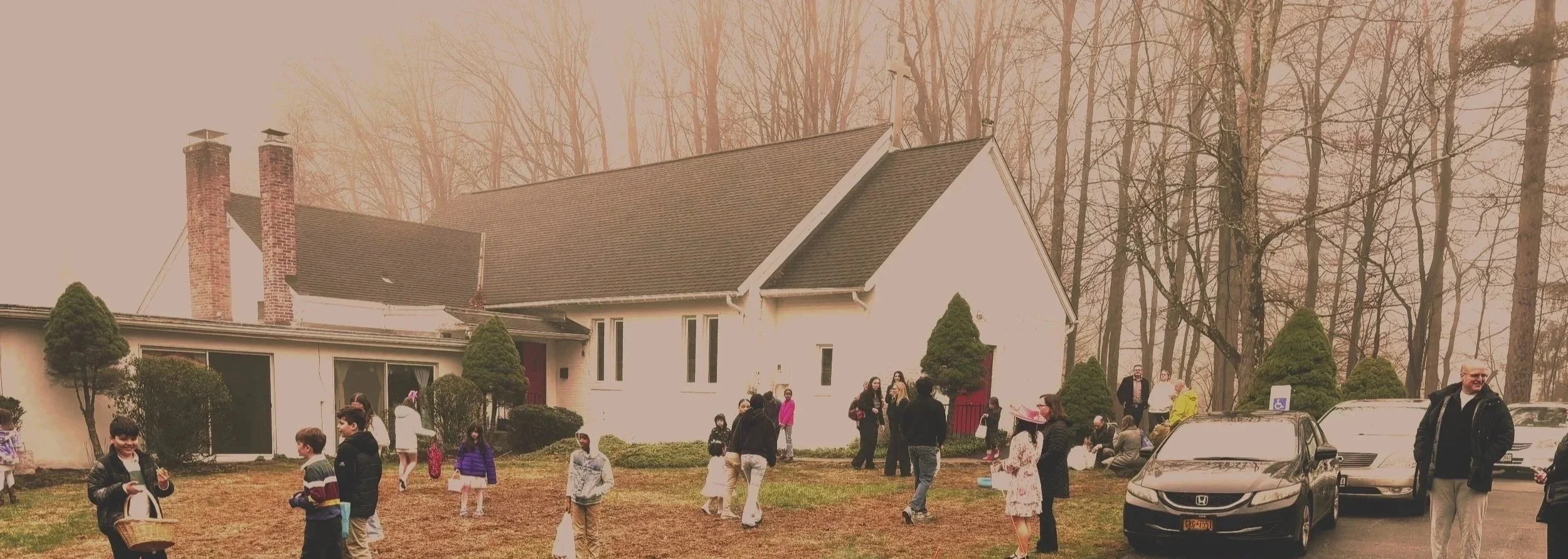 Group of children and adults gathered outside a white house with a sloped roof, surrounded by trees. Some children are holding baskets, and adults are standing or seated on chairs. Several cars are parked nearby.