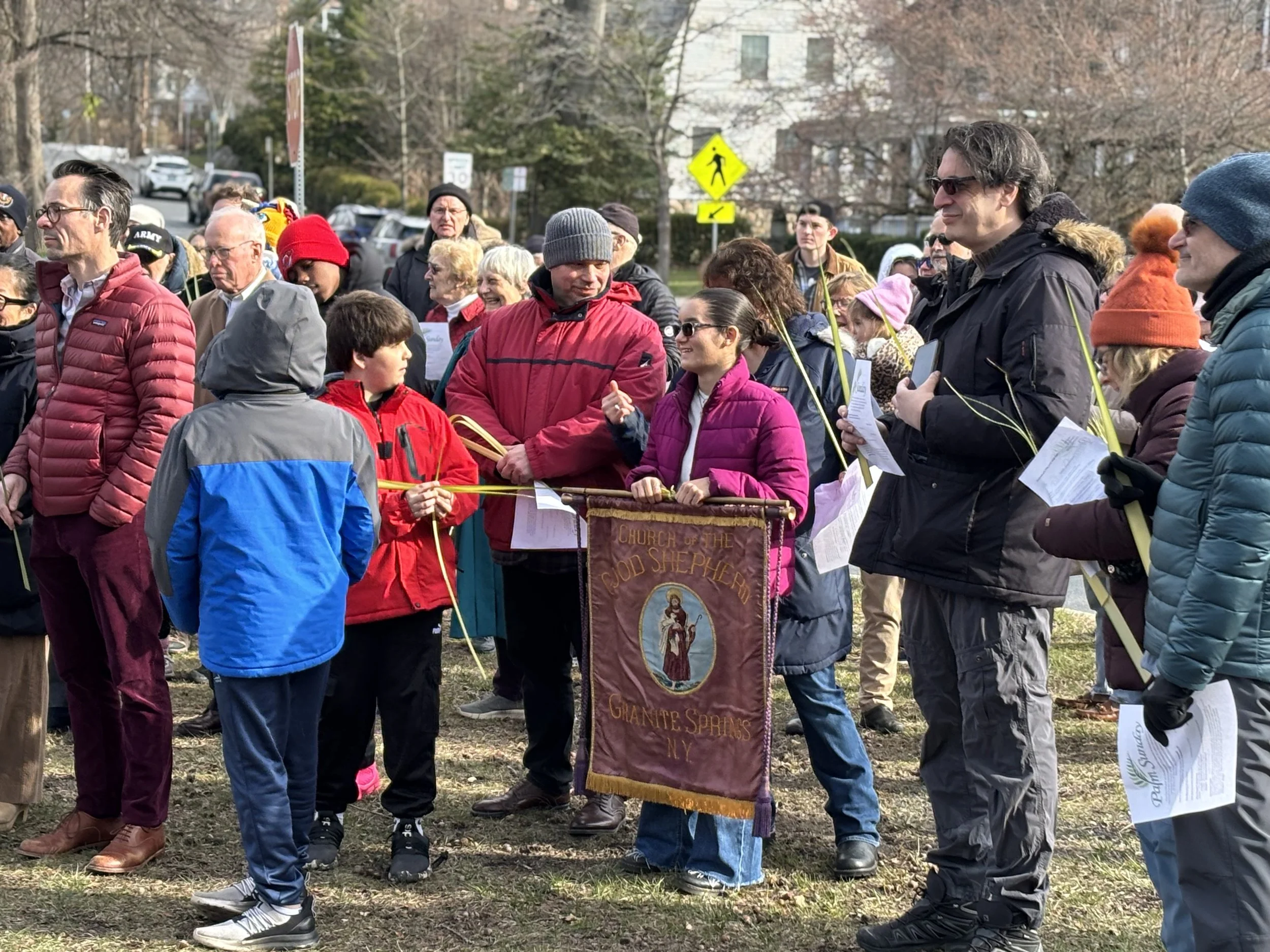 A diverse group of people gathered outdoors for a community event in winter, many wearing jackets, scarves, and hats. They are standing behind yellow rope barriers, holding papers, and some are smiling or talking. There is a banner with religious imagery and text, and a street sign in the background. The scene is set in a neighborhood with trees and houses.