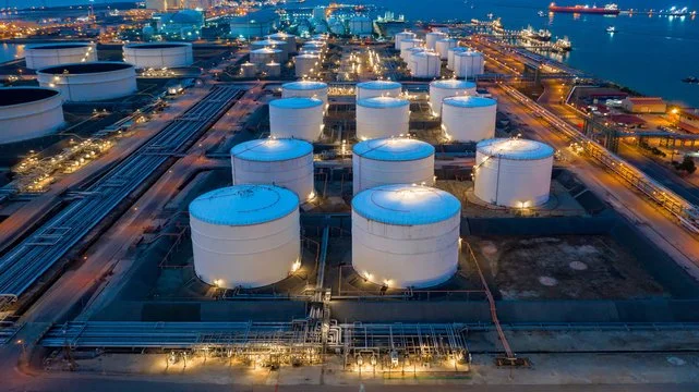 Aerial view of oil storage tanks at a port during nighttime, with city lights and ships visible in the background.
