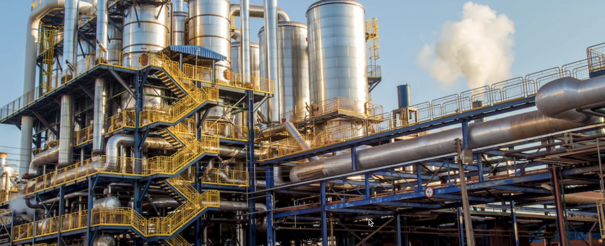 Industrial plant with multiple large silver tanks, yellow and blue staircases, and numerous silver pipes emitting steam against a blue sky.