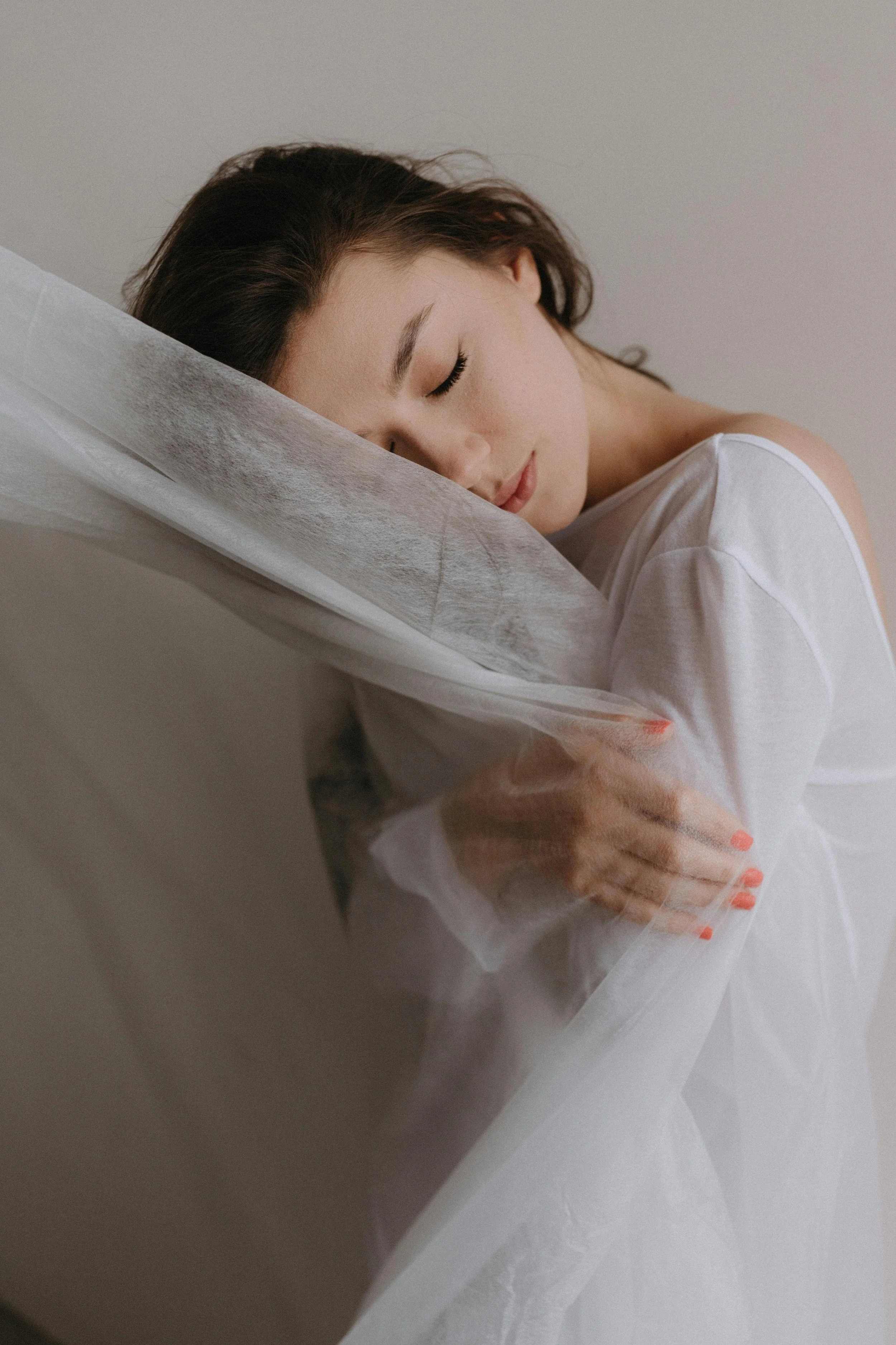 A woman with dark brown hair and light skin, resting her head on a pillow with her eyes closed. She is wearing a white shirt and appears to be sleeping or resting peacefully.