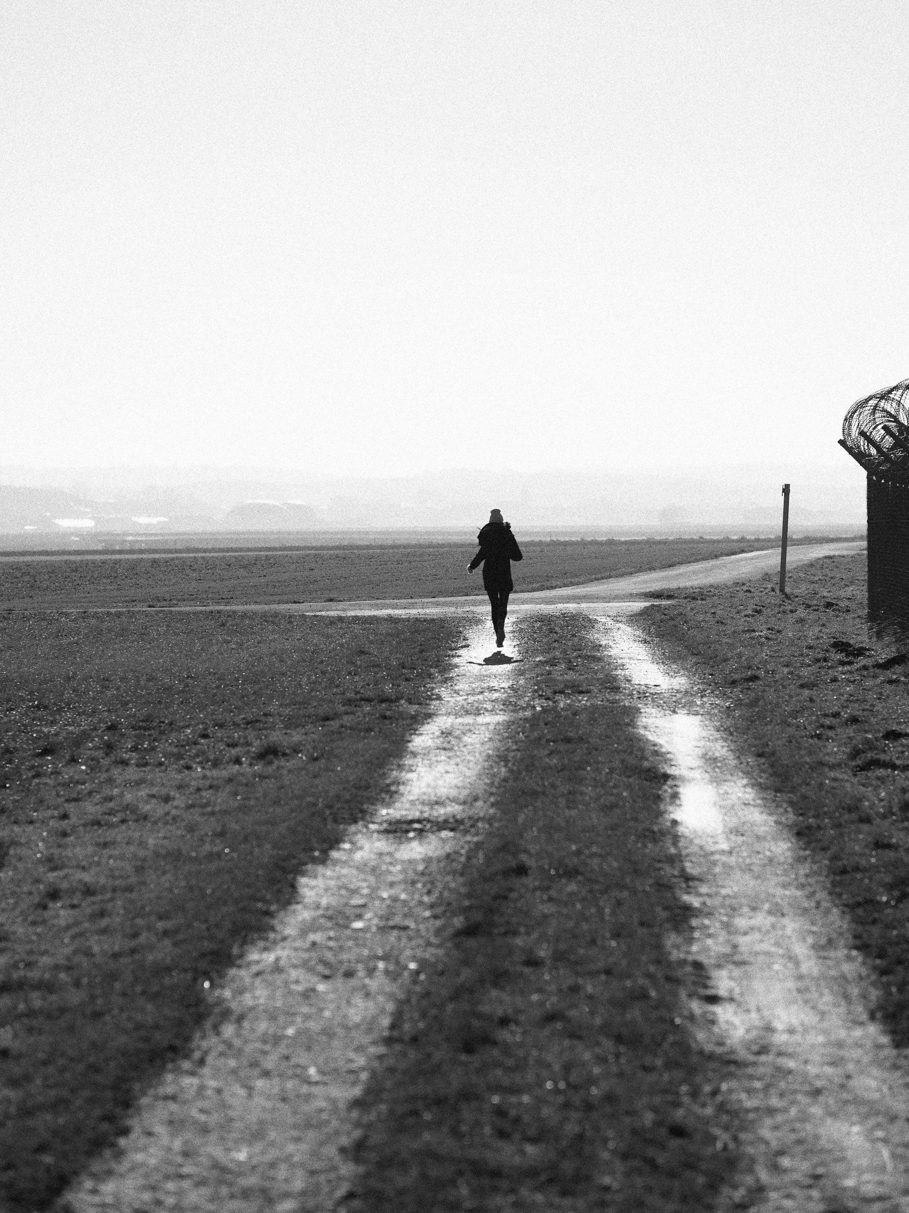 A person walking down a dirt path in a rural, open landscape with no buildings or trees visible in the distance. The photo is black and white, and the person is seen from behind.