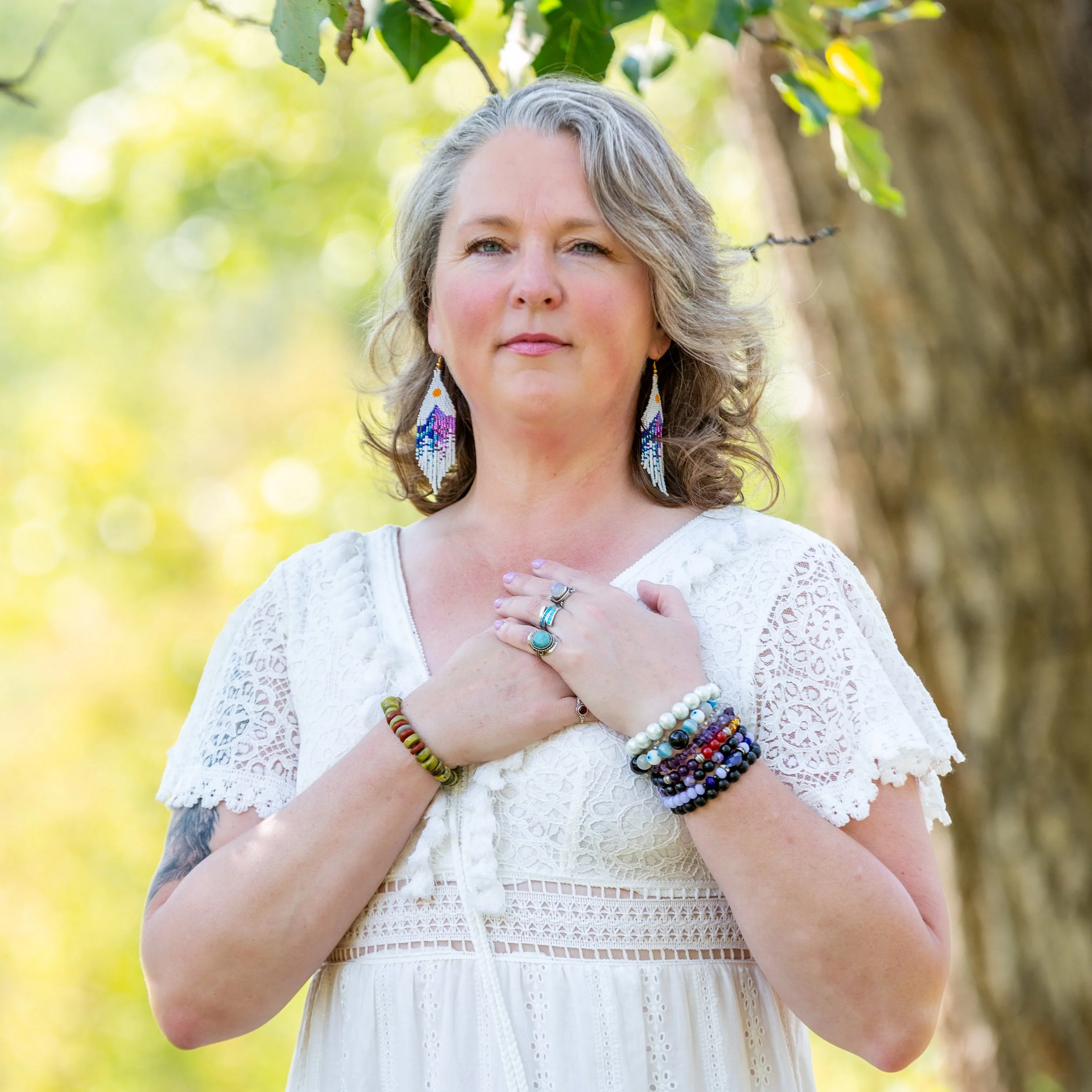 A woman with gray hair wearing a white lace dress and colorful beaded jewelry, standing outdoors with a blurred green background, holding her hand over her heart.
