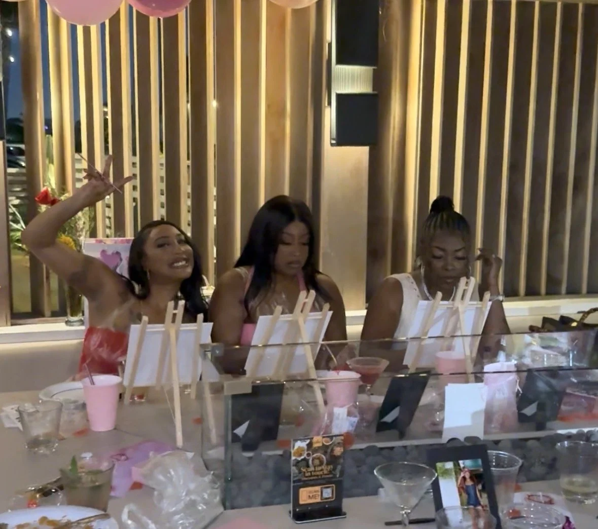 Three women sitting at a decorated table with pink cups and photo props, enjoying a celebration at a restaurant.