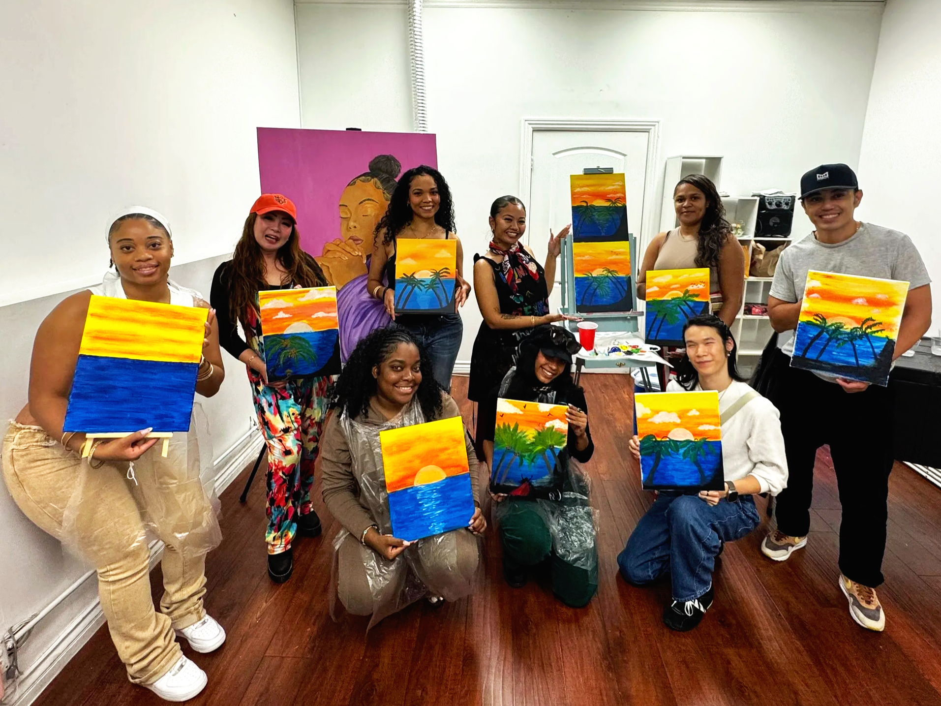 Group of people holding up paintings of a colorful sunset over the ocean, after a painting class in a room with white walls and wooden flooring.