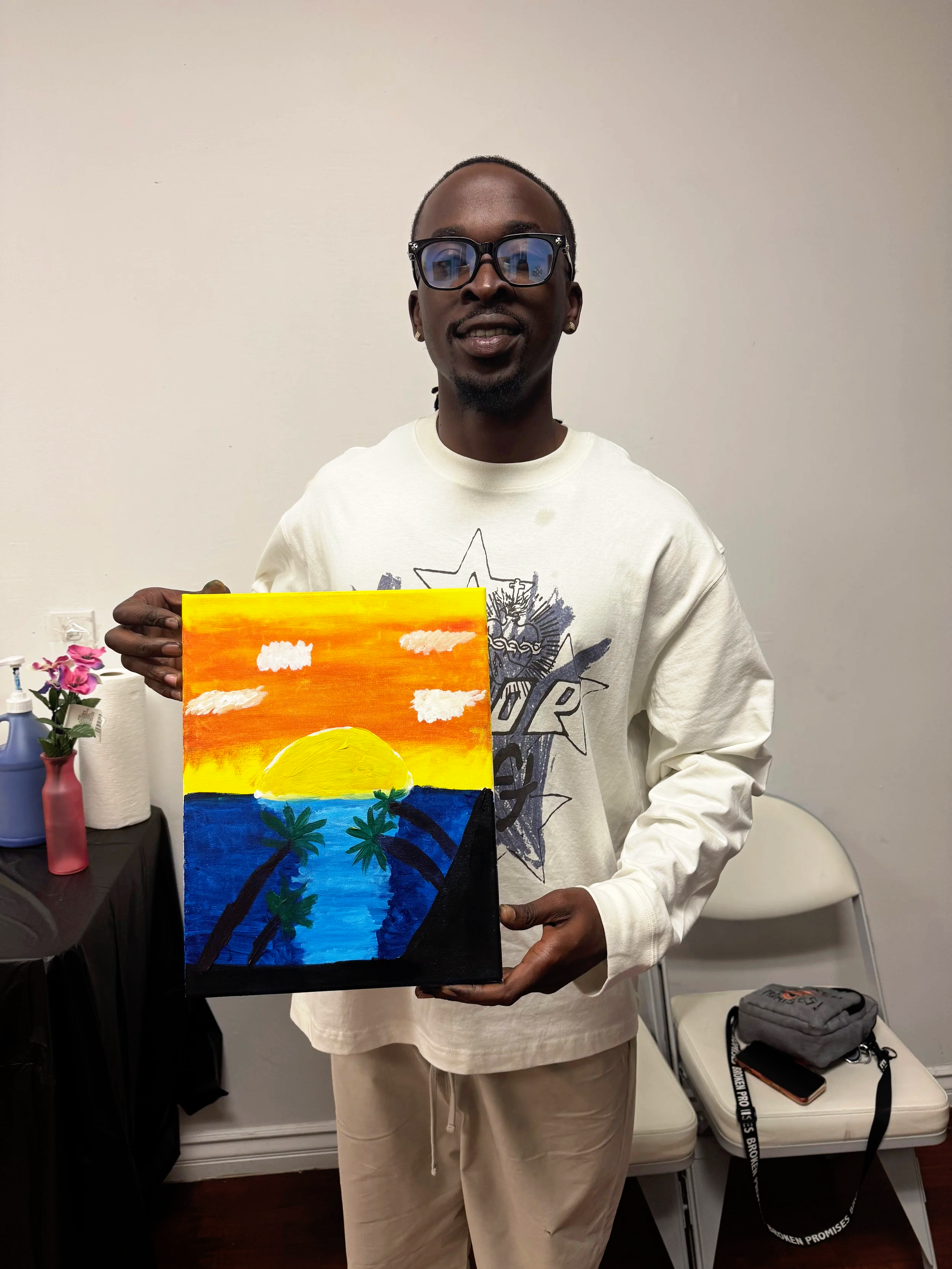 A man holding a colorful painting of a sunset over the ocean with palm trees in the foreground.