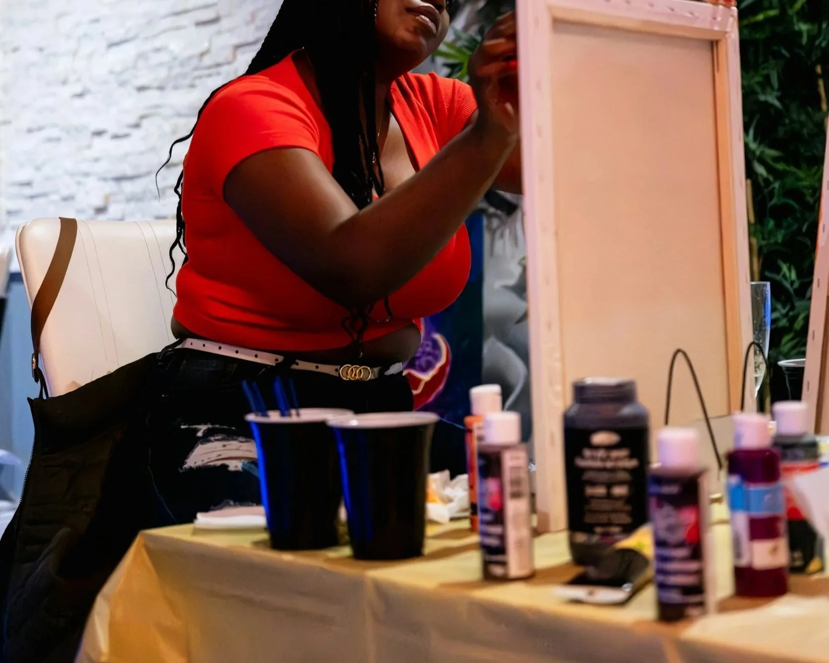 A woman in a red shirt sitting at a table with various bottles of paint and art supplies, working on a small canvas or paper. She is focused on her artwork, with a background of a white brick wall and green foliage.