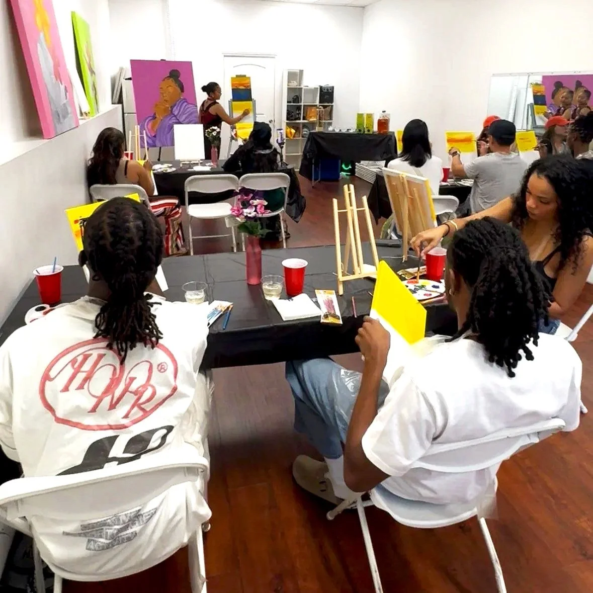 People participating in a group painting class in a brightly lit studio, with canvas and art supplies on tables and artwork displayed on the walls.