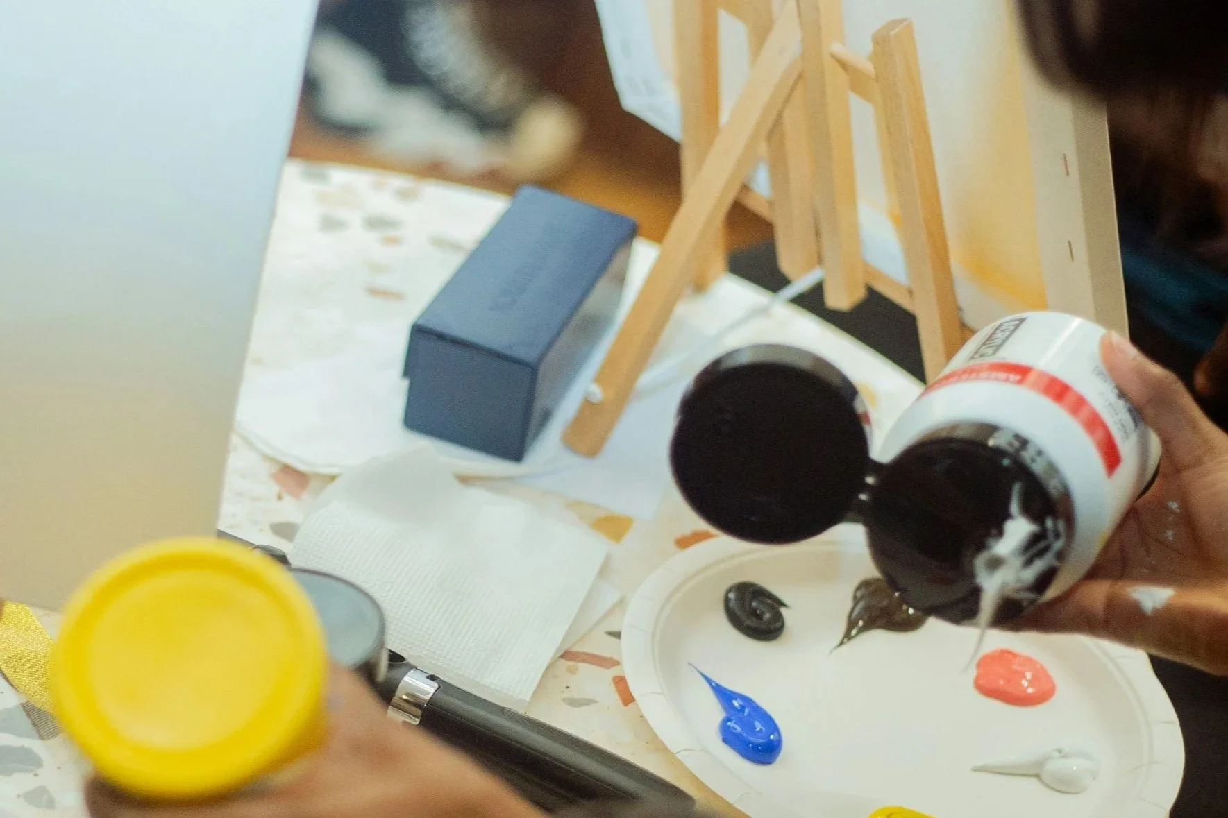A person pouring black paint from a white bottle with red label onto a palette with blue, black, orange, and white paint, in front of a laptop and small wooden easel.