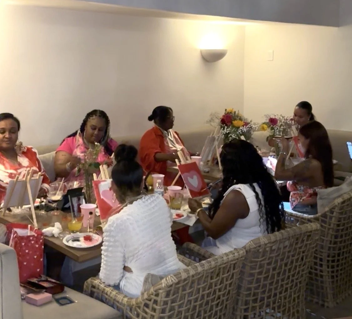 Women gathered at a table engaged in a painting activity, surrounded by art supplies, drinks, and flowers, during a social event.
