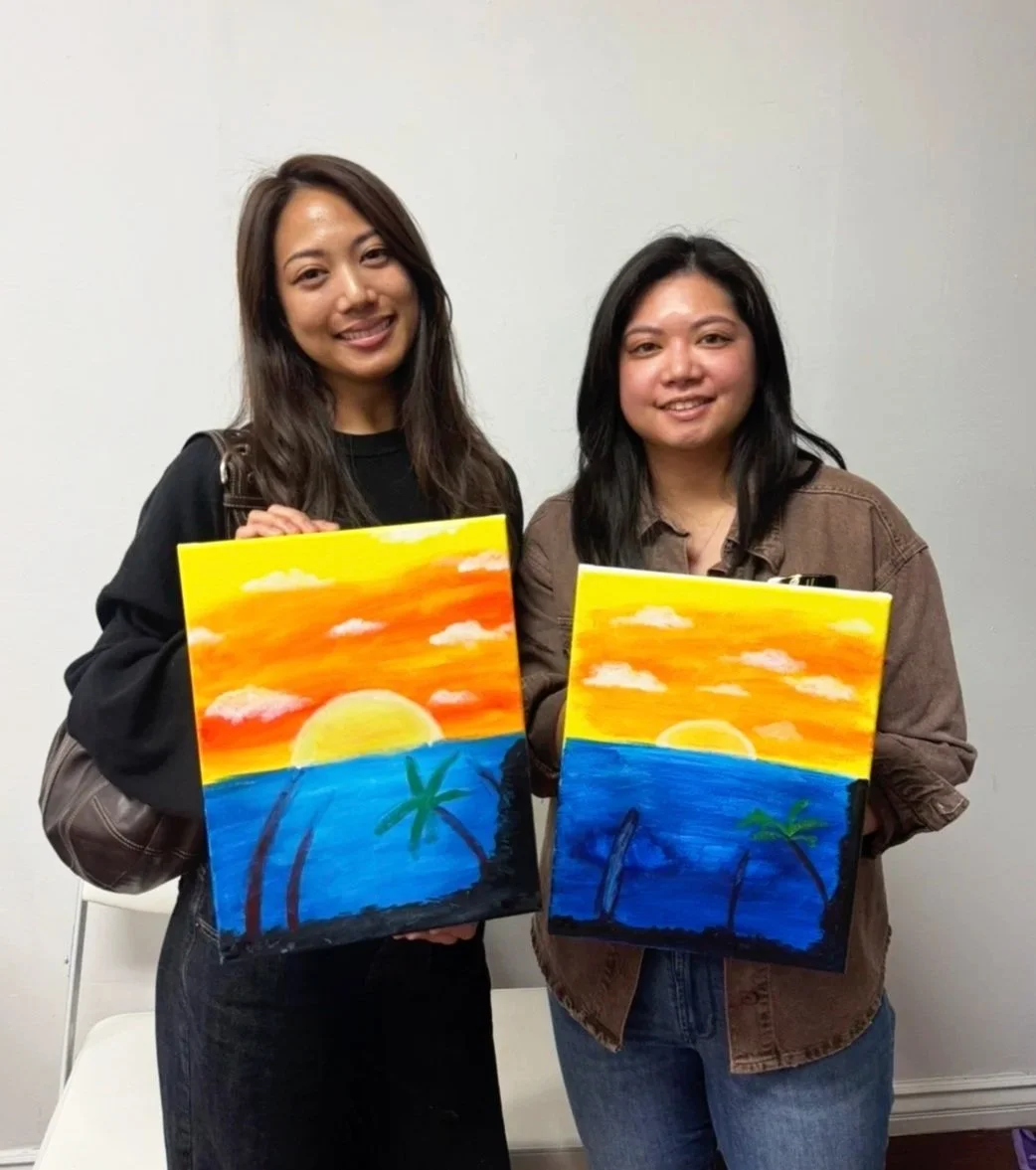 Two women standing indoors holding paintings of a sunset over the ocean with palm trees.
