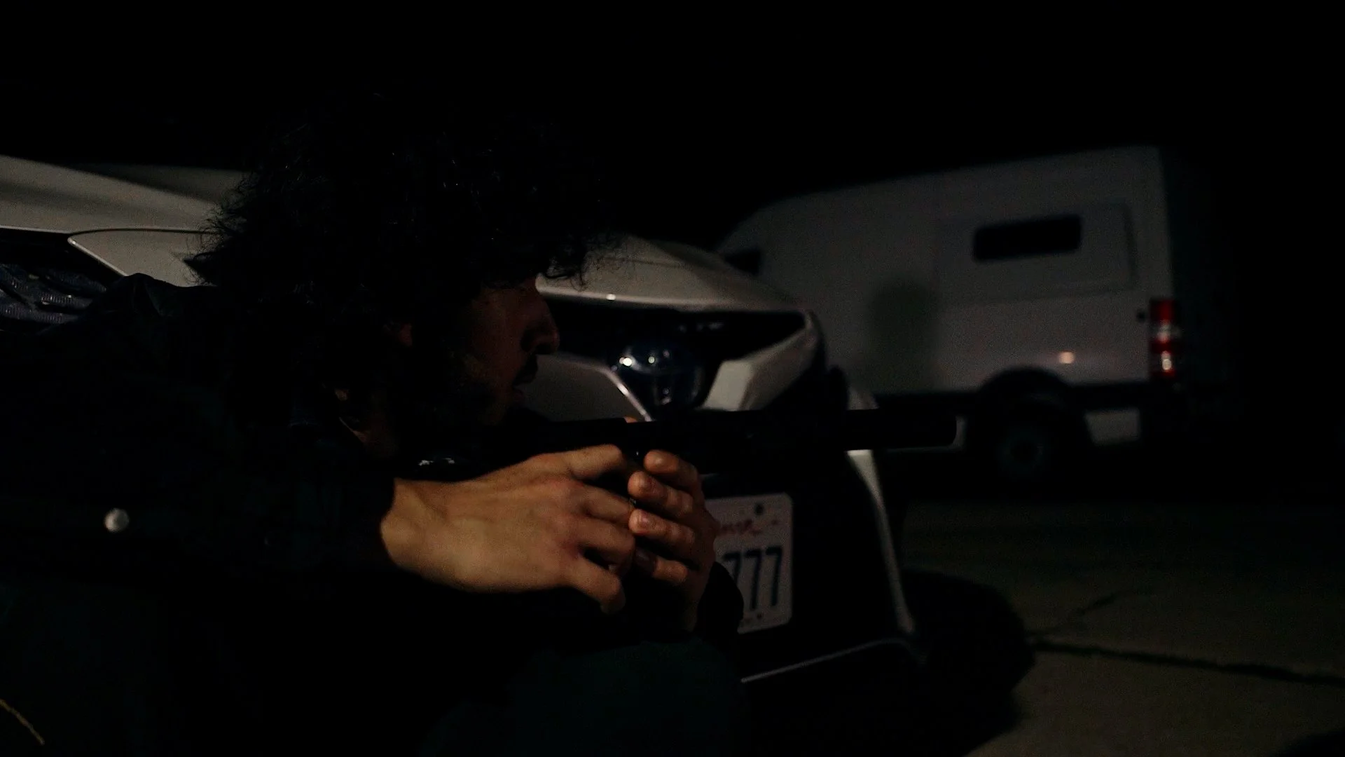 Person with dark hair crouching and holding a gun in a dark environment with vehicles in the background and a shadowy figure approaching.