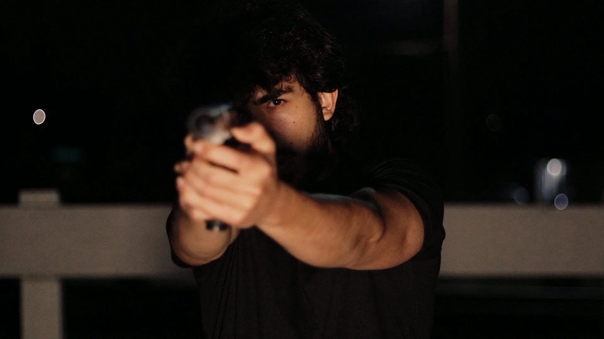 A young man with dark hair and a beard, aiming a gun with one eye closed against a dark background.