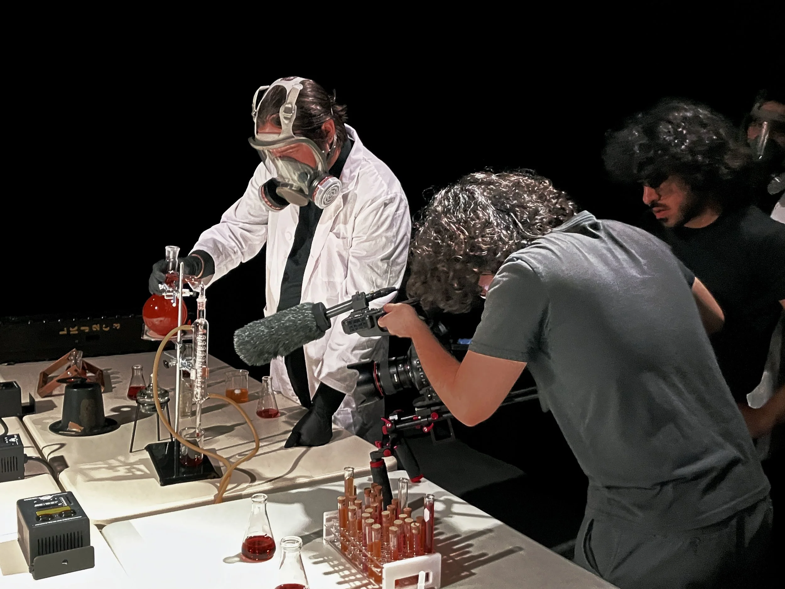 Behind-the-scenes of a director filming a scene of scientists conducting experiments at a laboratory table with glassware and electronic equipment, wearing goggles and lab coats, dark background, cinematic lighting, film set atmosphere.