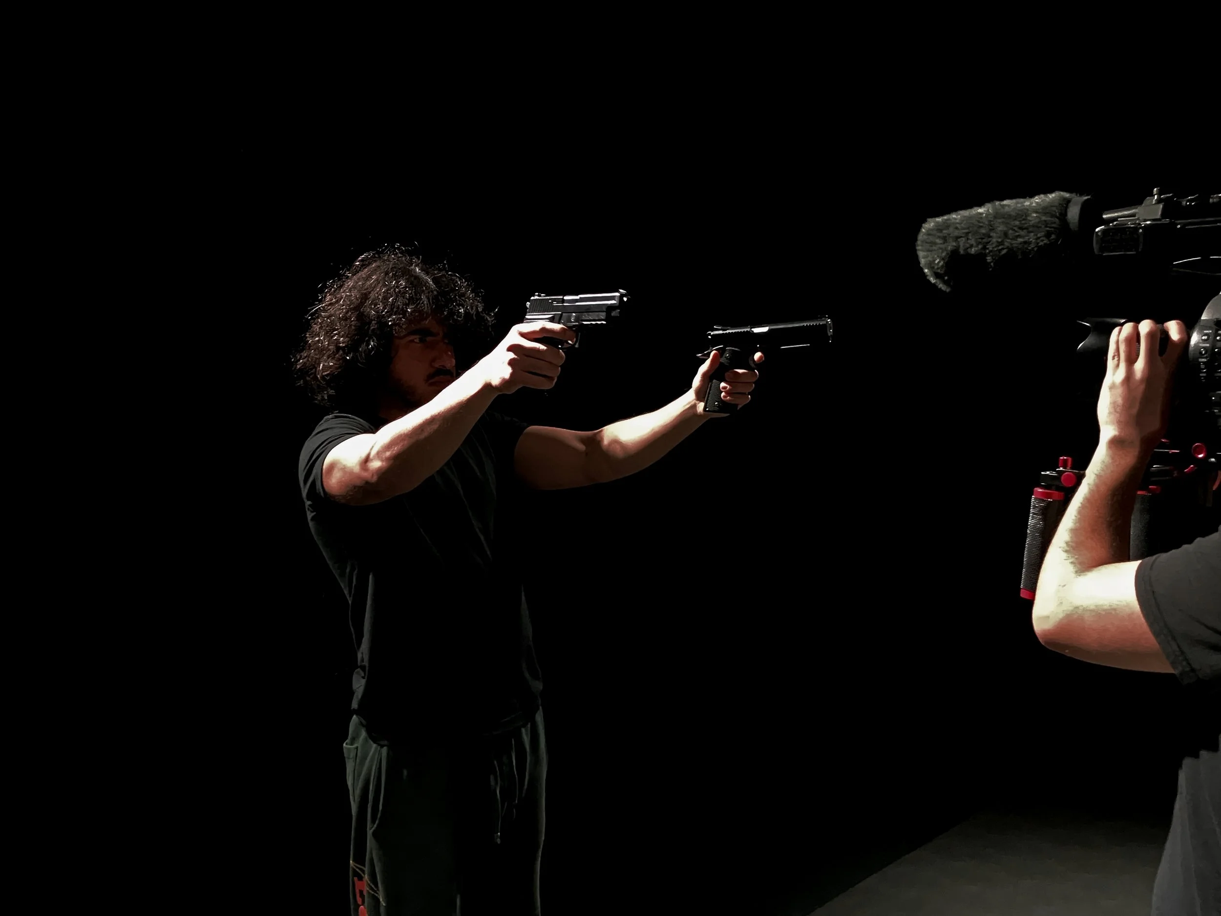 A cinematic behind-the-scenes shot of a director-actor aiming two handguns, with a crew member filming them up close using a camera, set against a black background, dramatic lighting, film set atmosphere, realistic style.