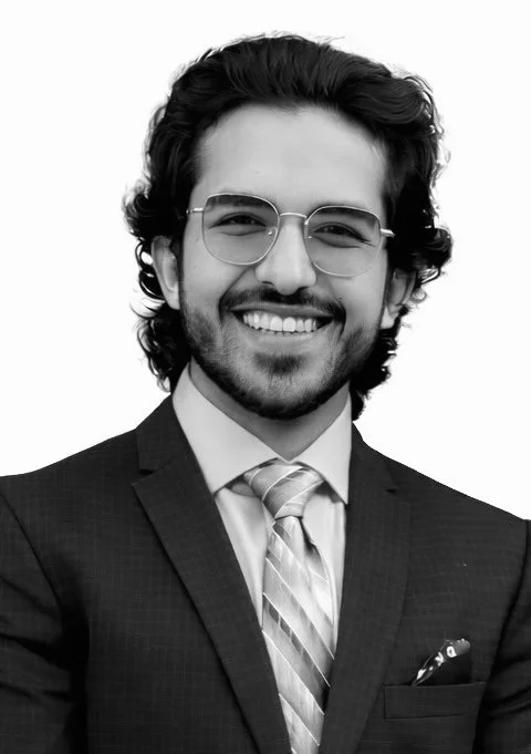 Black-and-white director headshot of a man with dark wavy hair, glasses, and a beard, wearing a suit and tie, smiling, clean studio lighting, professional and cinematic portrait style.