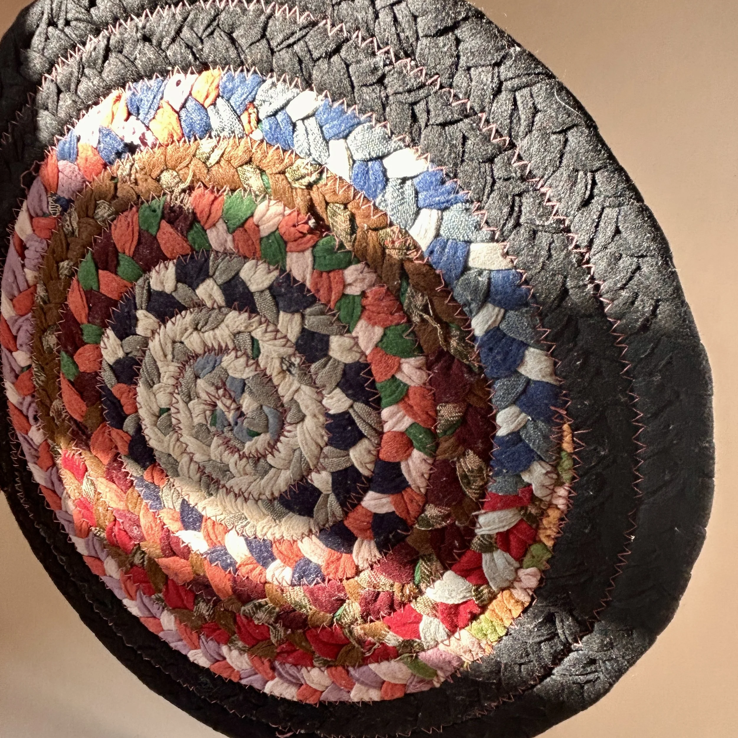 Vintage round braided fabric table mat made from multicolor textile scraps in concentric rings with a wide black braided border and zigzag stitching.