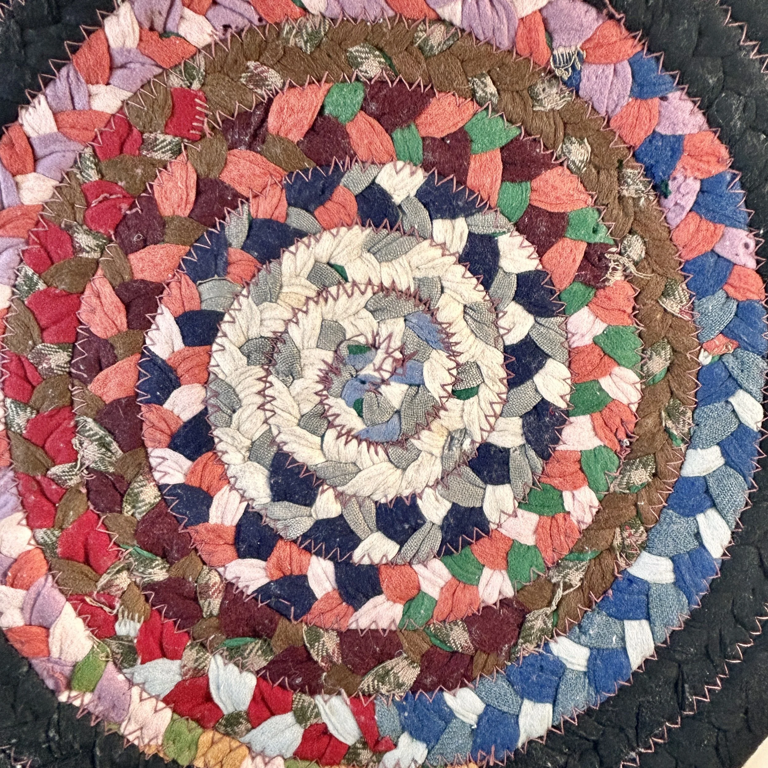 Vintage round braided fabric table mat made from multicolor textile scraps in concentric rings with a wide black braided border and zigzag stitching.