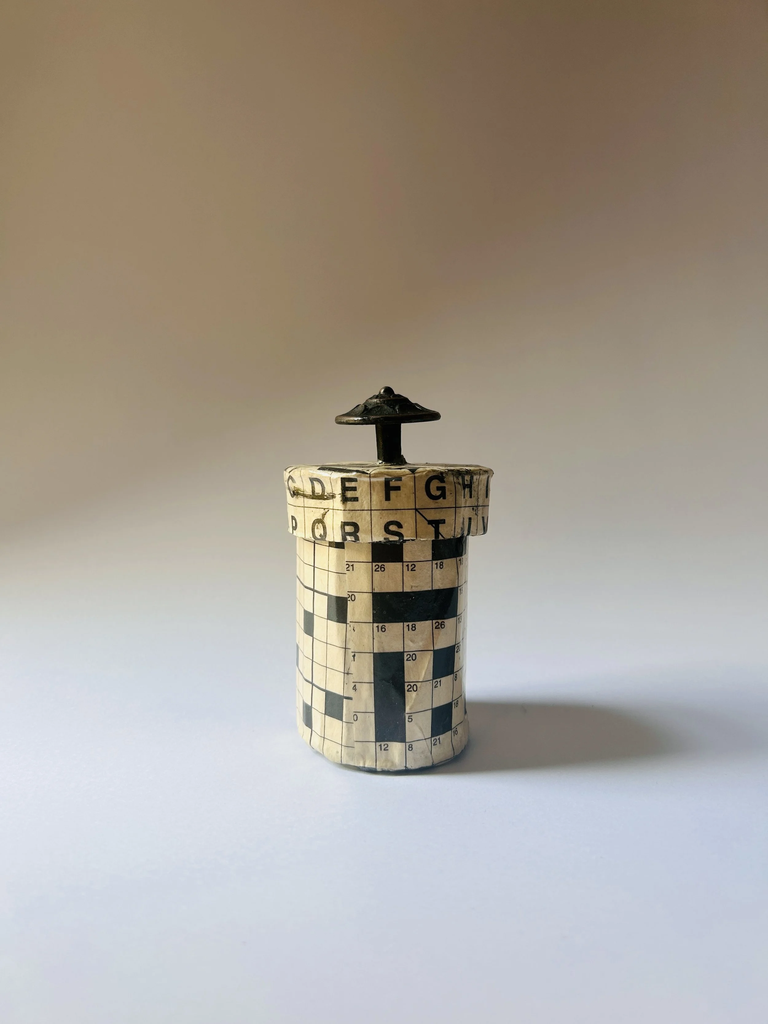 Vintage lidded box covered in crossword puzzle print with dark sculptural knob, graphic paper collage object