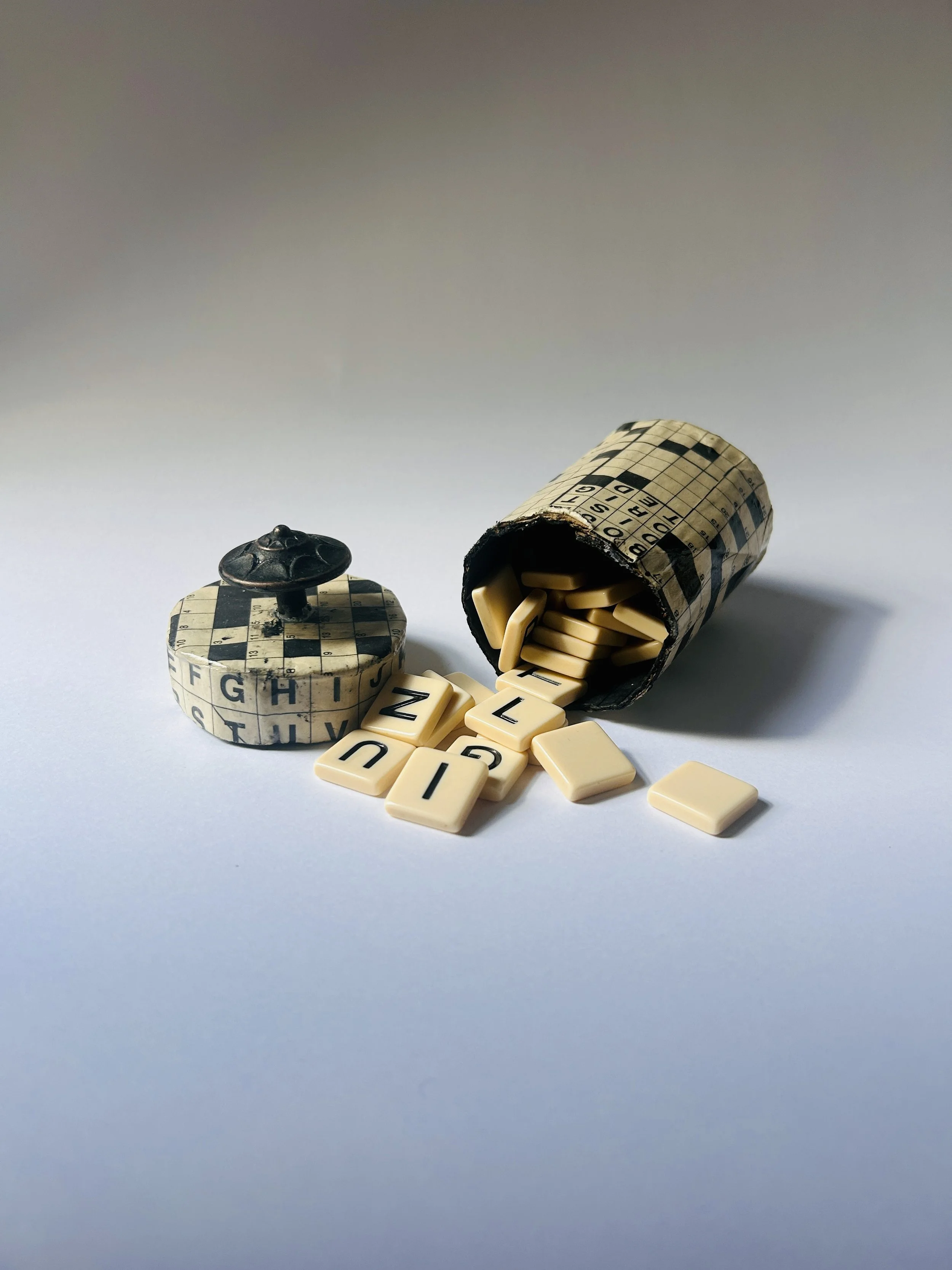 Vintage lidded box covered in crossword puzzle print with dark sculptural knob, graphic paper collage object
