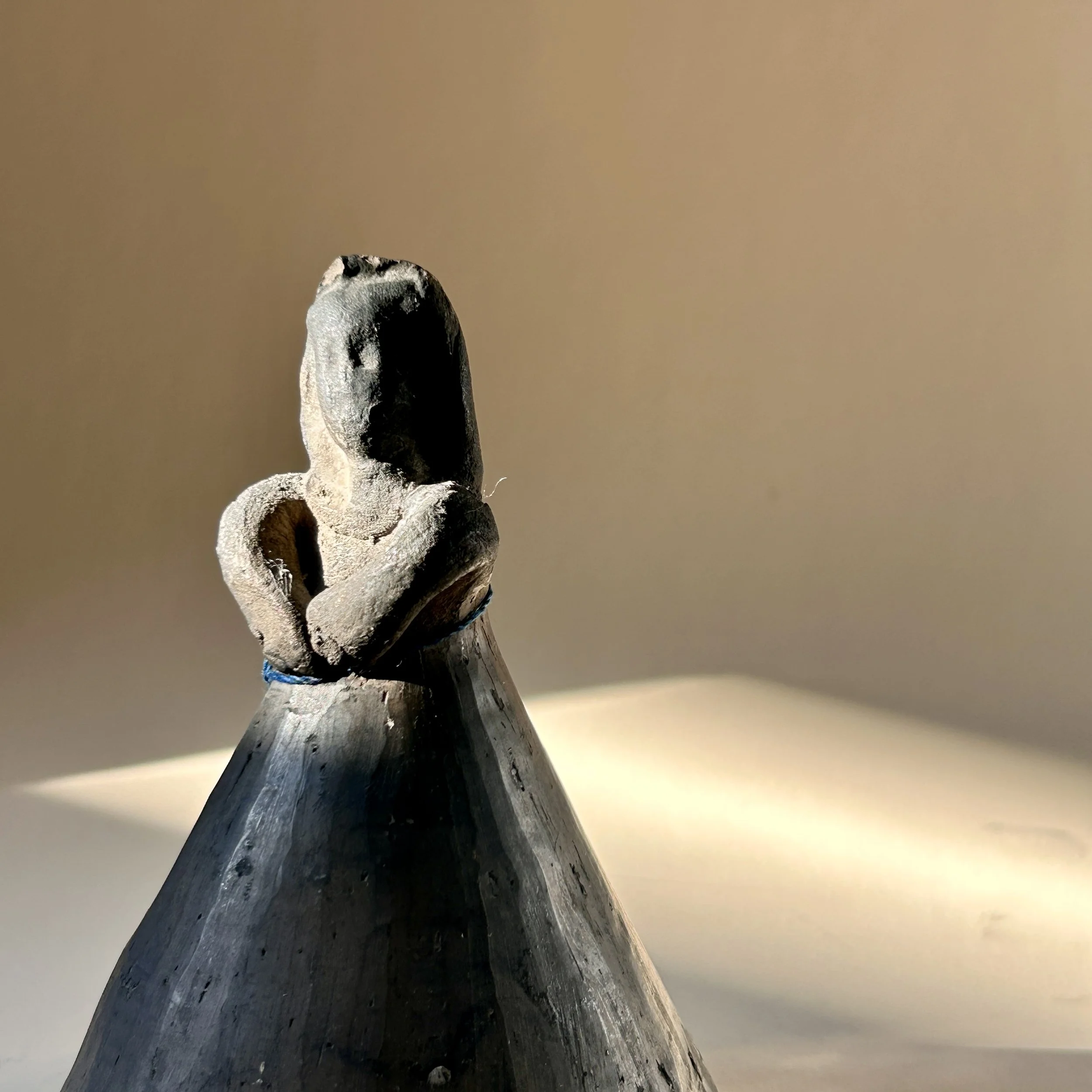 Vintage handmade black clay angel bell with folded arms, wide bell-shaped skirt and small clay clapper suspended by blue cord.