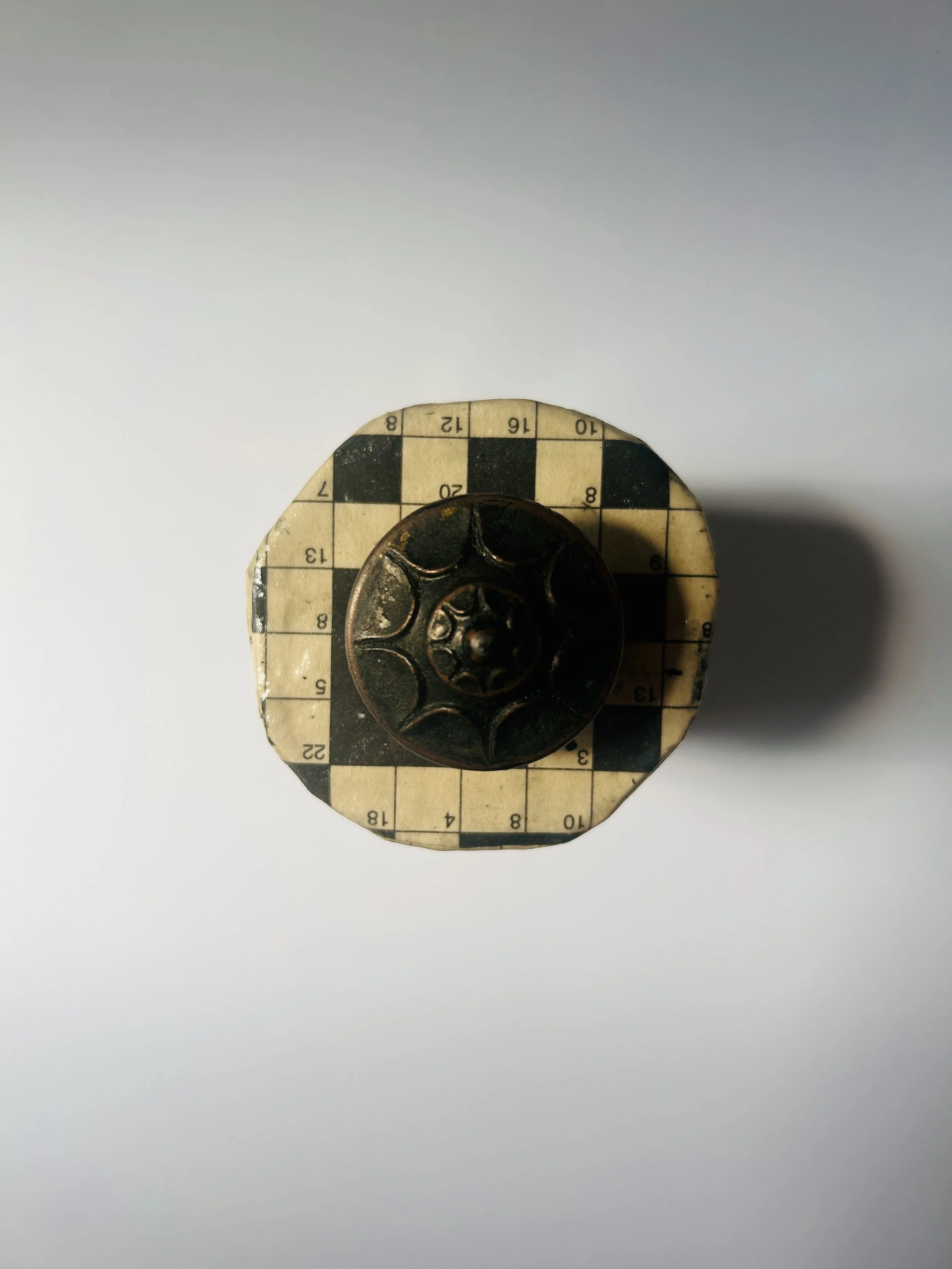 Vintage lidded box covered in crossword puzzle print with dark sculptural knob, graphic paper collage object