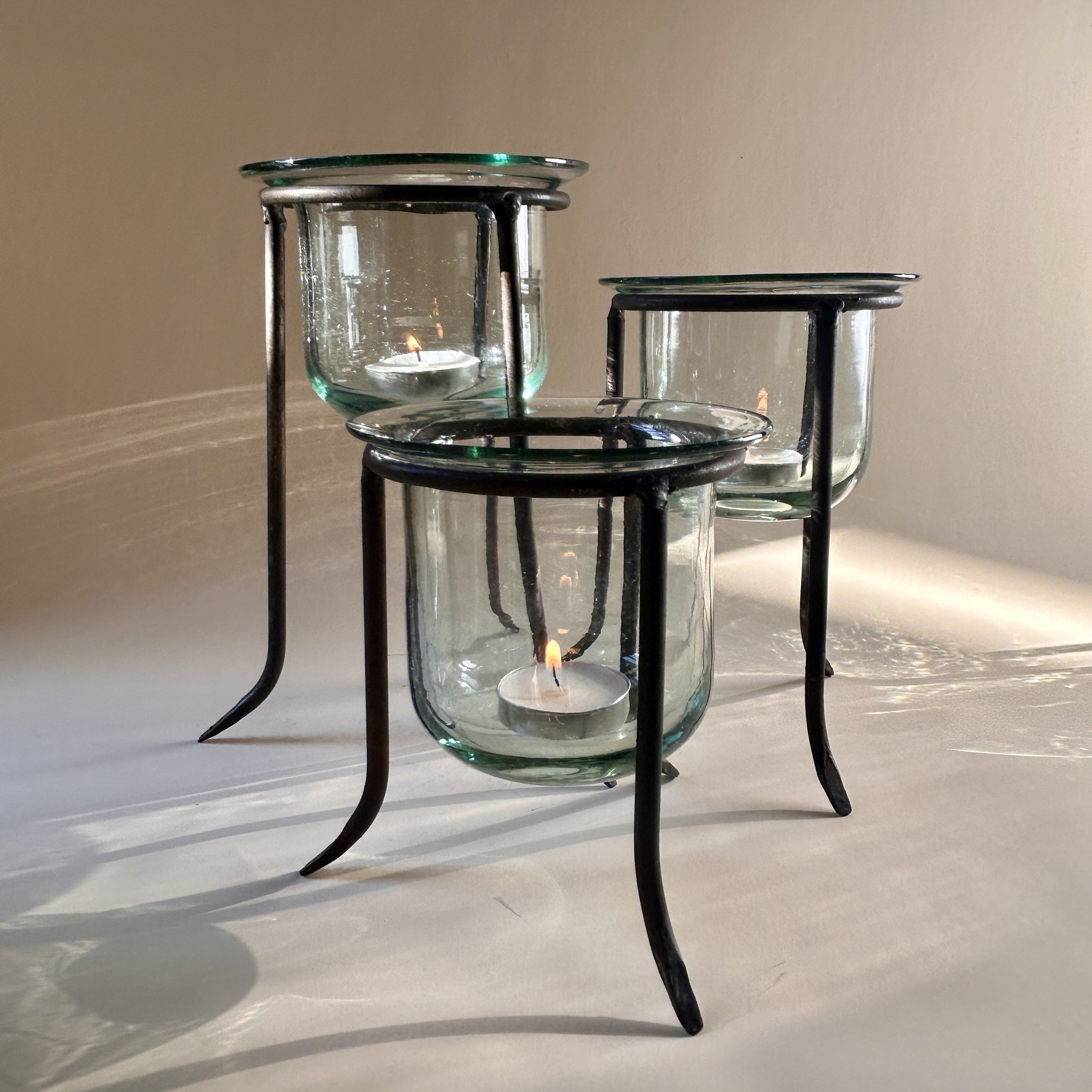 Set of three clear handblown glass tealight holders in dark iron tripod stands with curved legs, shown in graduated heights.