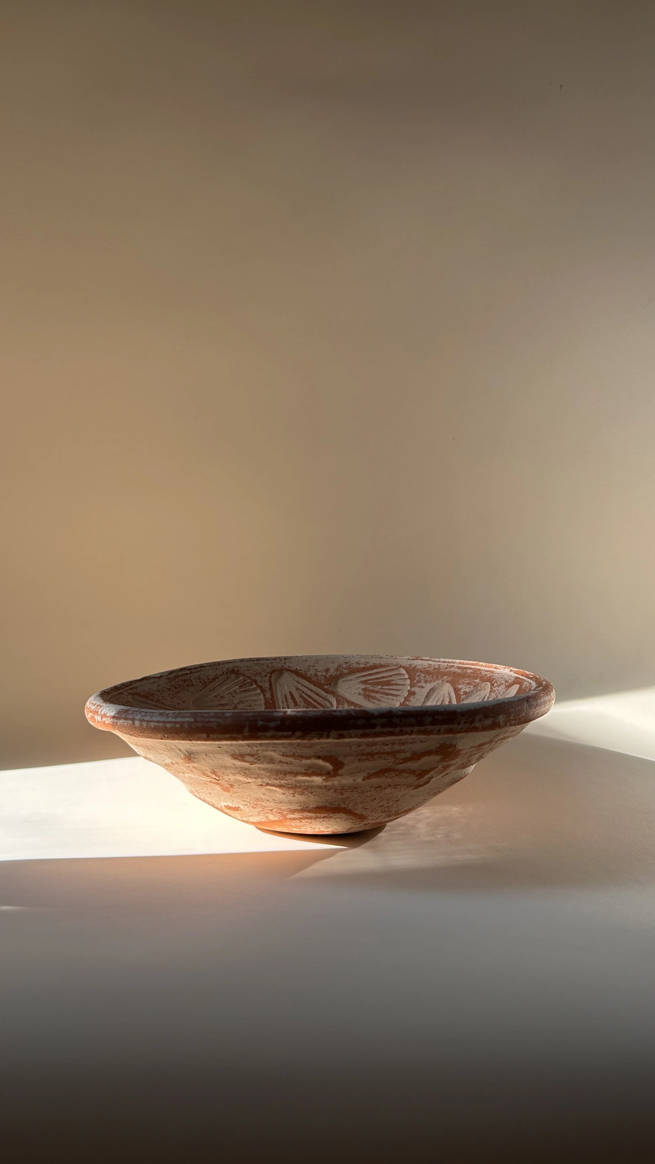 Shallow terracotta pottery bowl with raised scallop shell motifs and distressed chalky white finish over warm red clay.