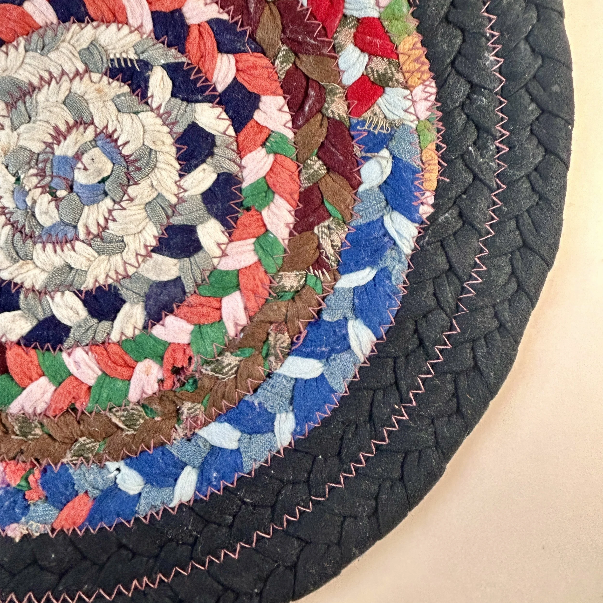 Vintage round braided fabric table mat made from multicolor textile scraps in concentric rings with a wide black braided border and zigzag stitching.
