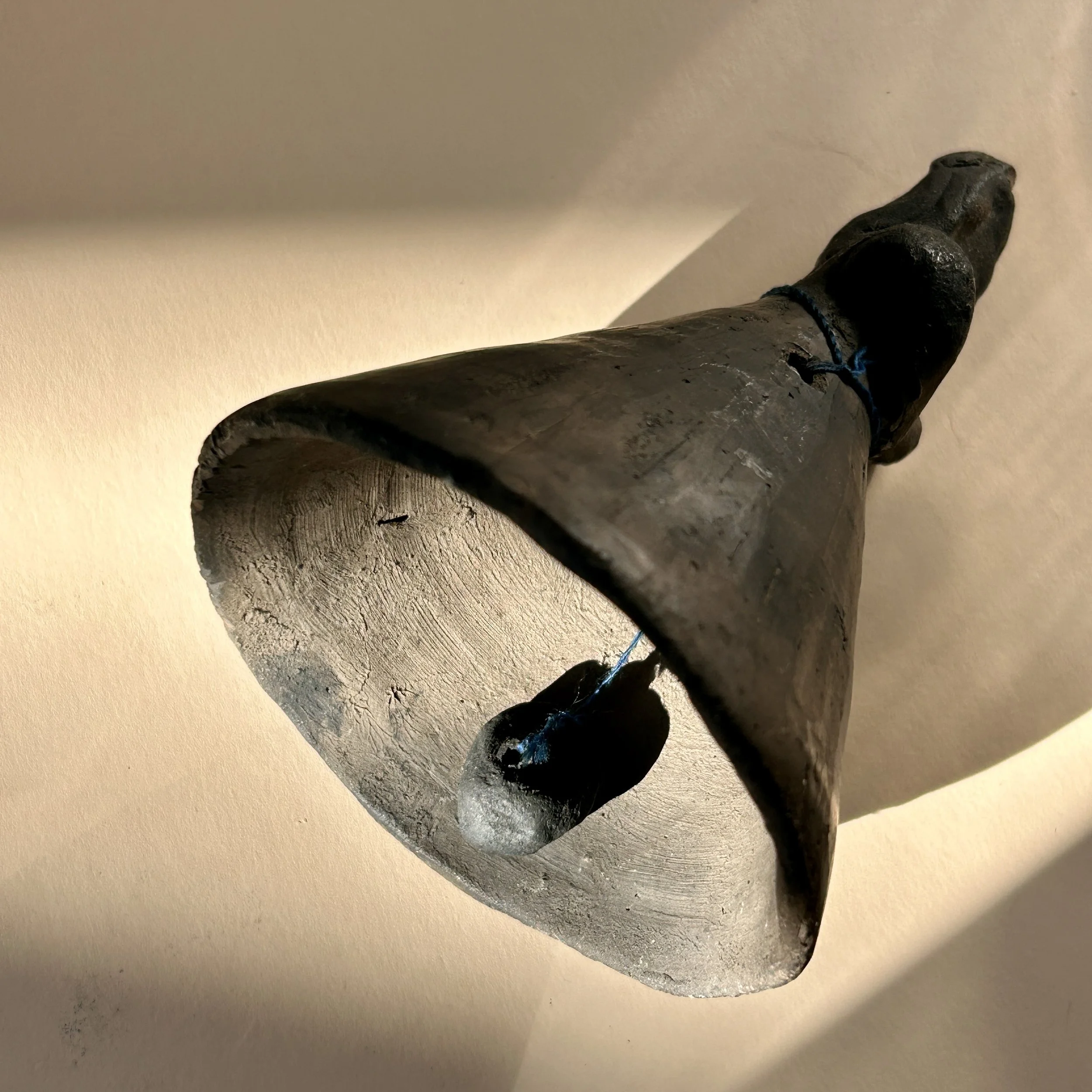 Vintage handmade black clay angel bell with folded arms, wide bell-shaped skirt and small clay clapper suspended by blue cord.
