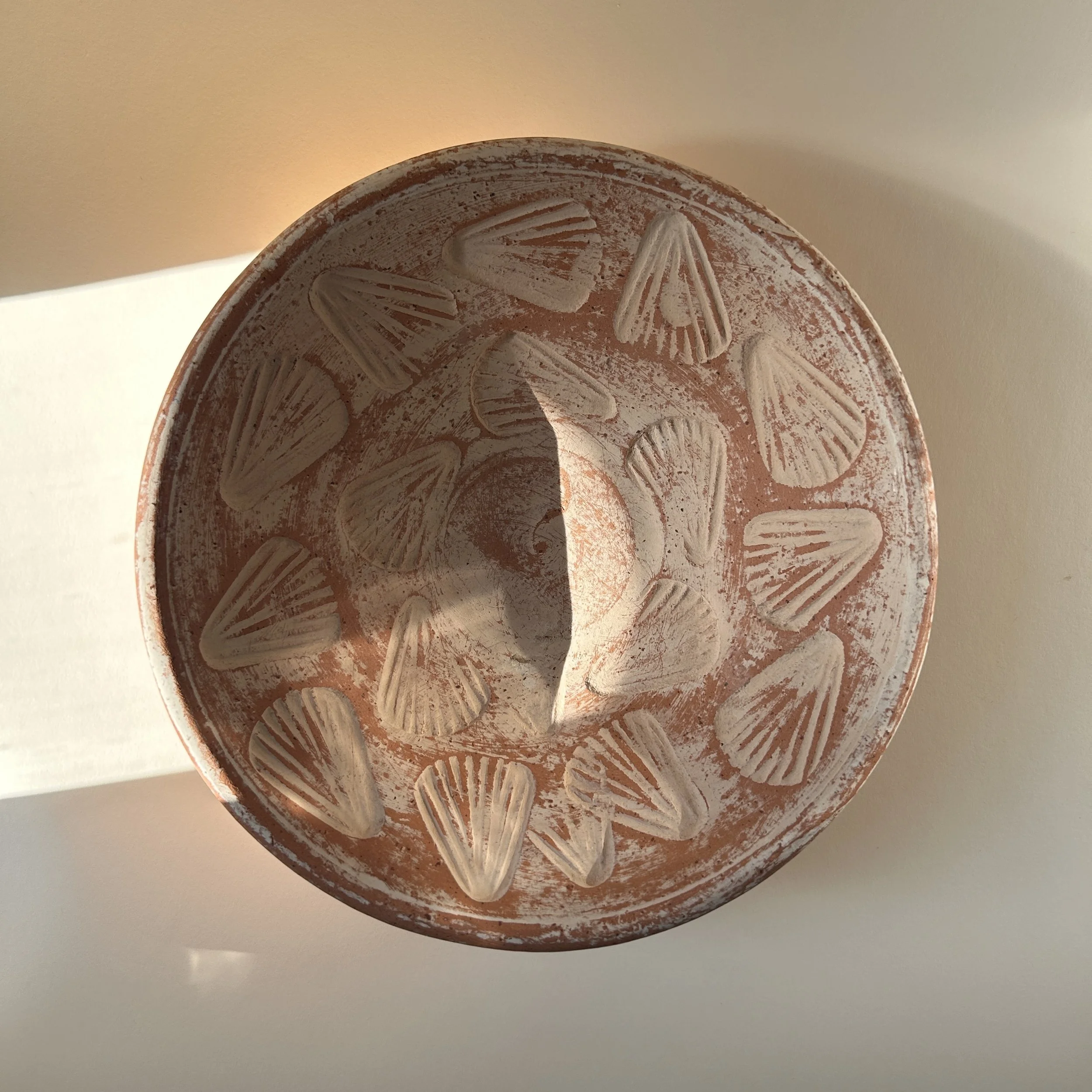 Shallow terracotta pottery bowl with raised scallop shell motifs and distressed chalky white finish over warm red clay.