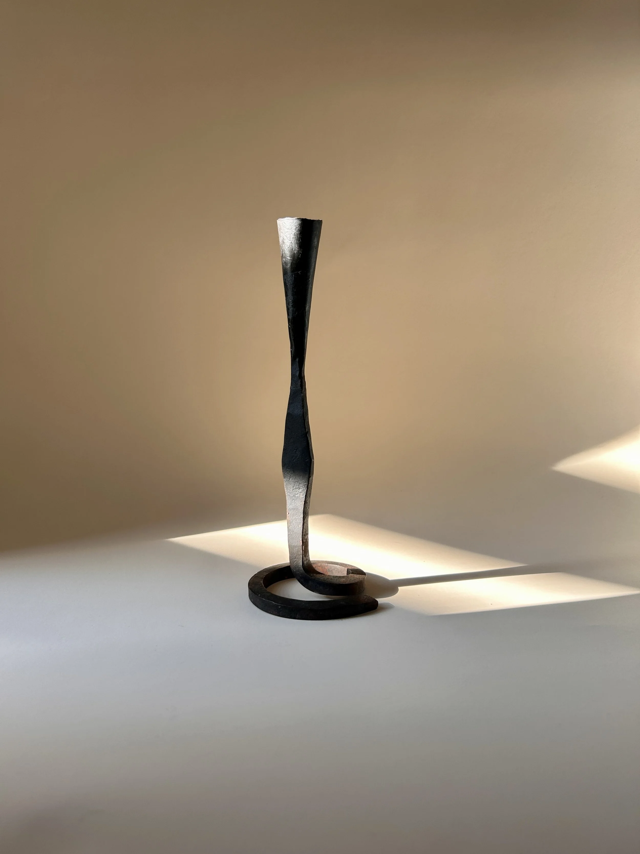 Sculptural hand forged black iron taper candlestick with a curled spiral base, slender twisted stem, and flared candle cup.