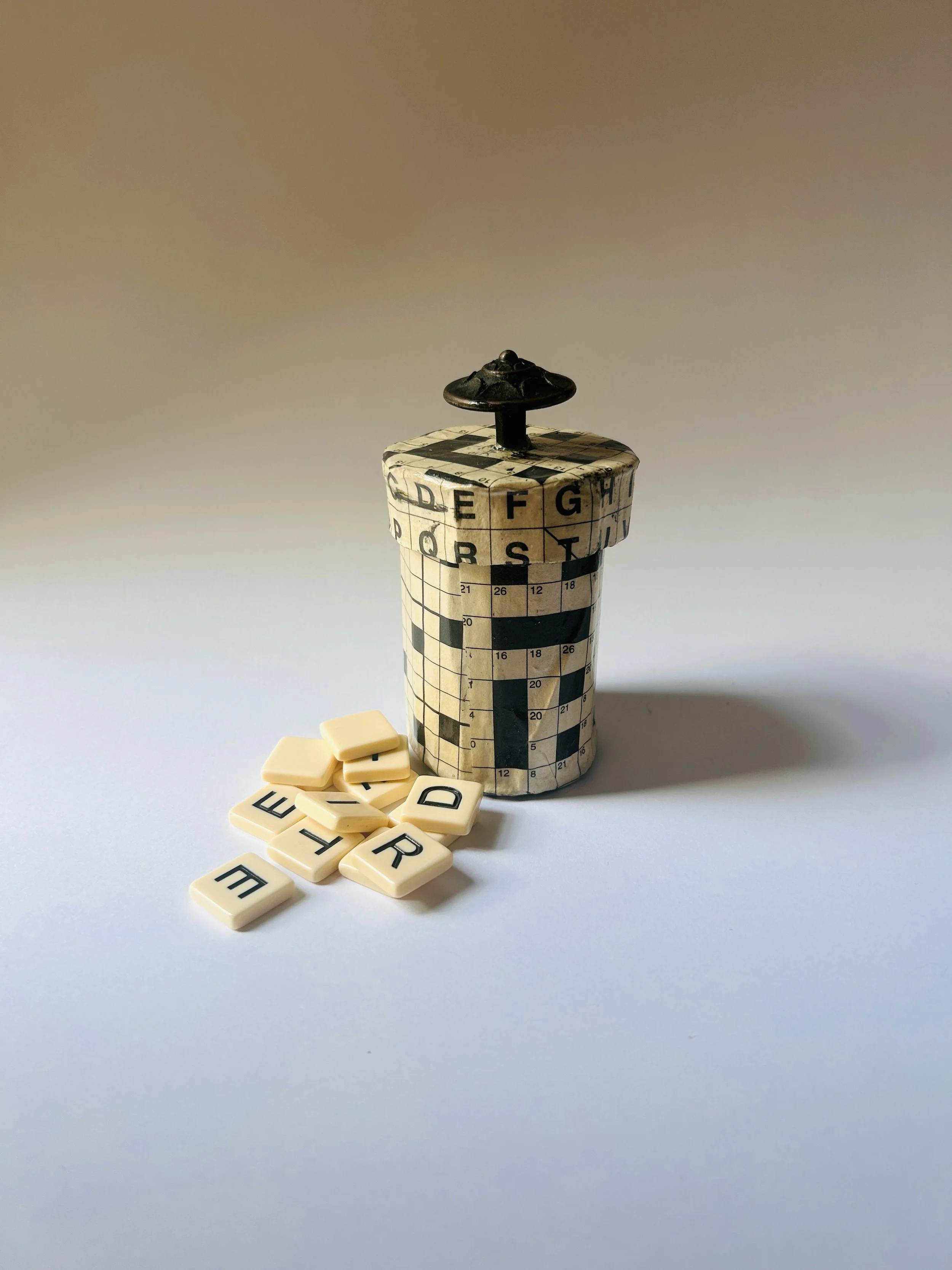 Vintage lidded box covered in crossword puzzle print with dark sculptural knob, graphic paper collage object