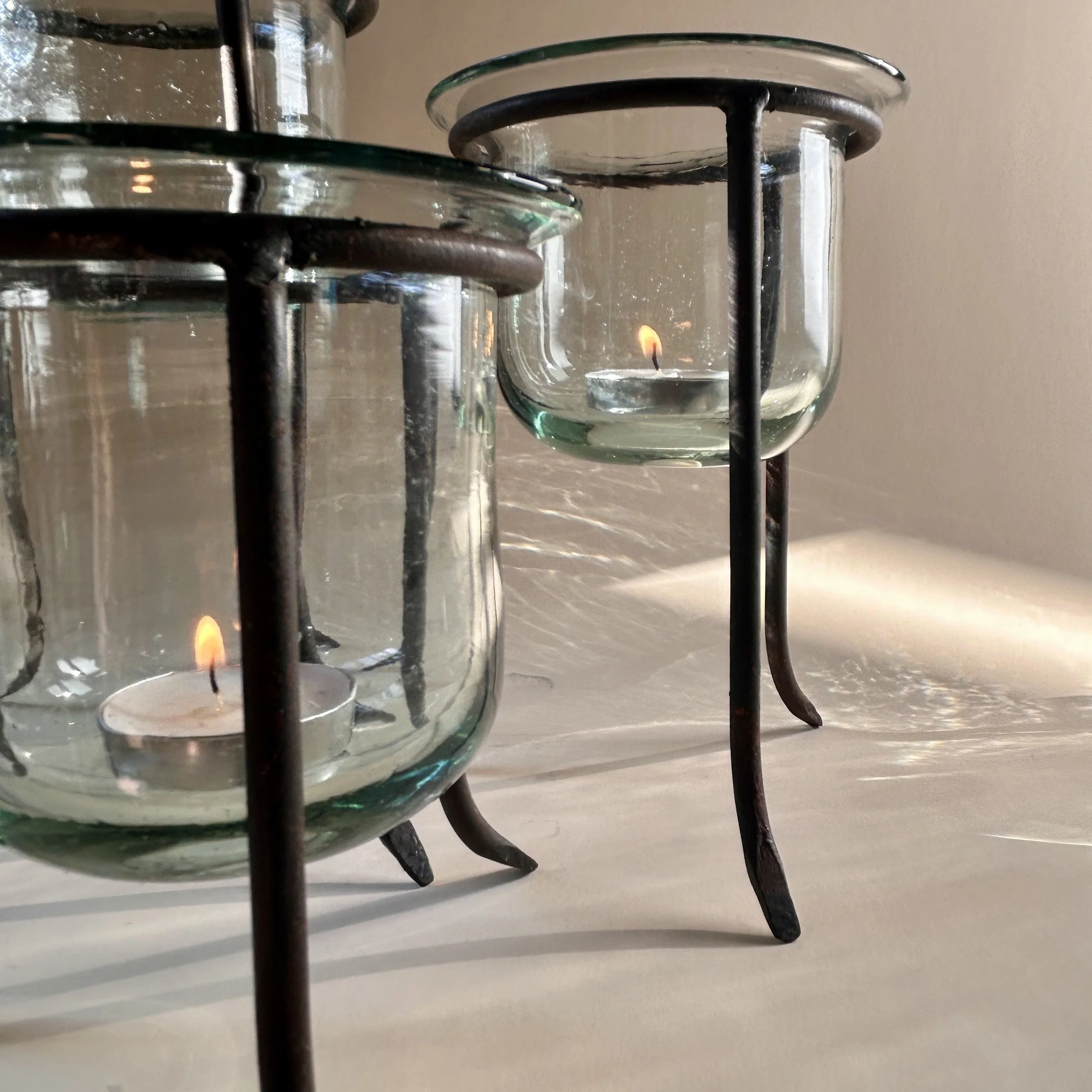 Set of three clear handblown glass tealight holders in dark iron tripod stands with curved legs, shown in graduated heights.