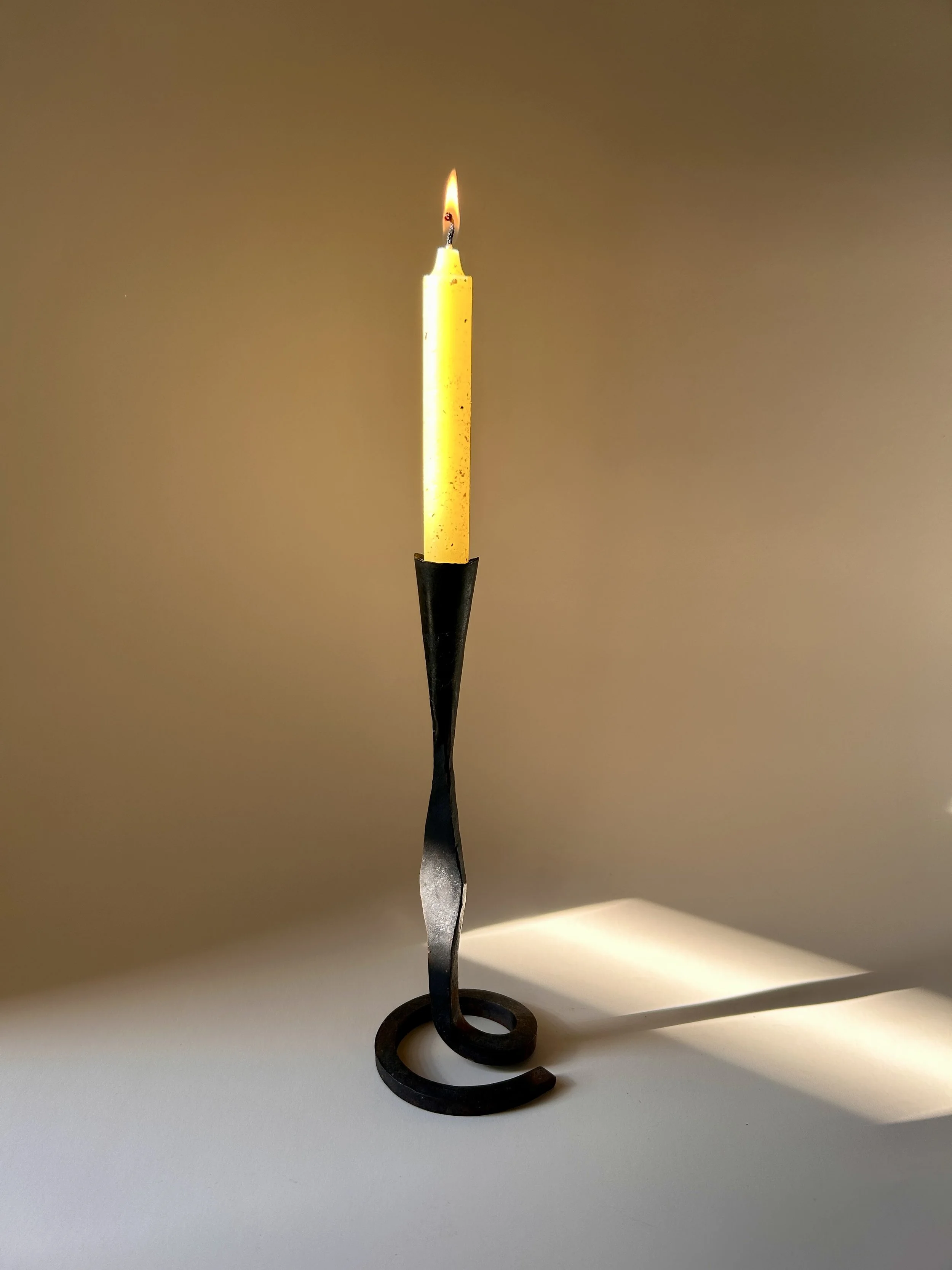 Sculptural hand forged black iron taper candlestick with a curled spiral base, slender twisted stem, and flared candle cup.