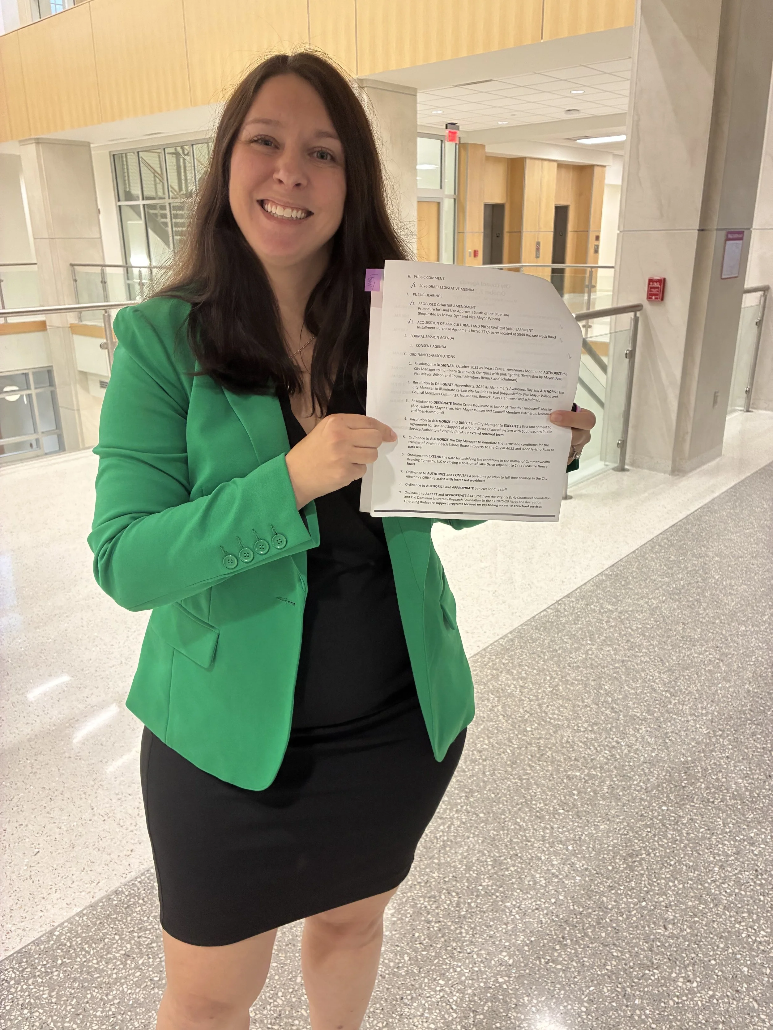 A woman with long dark hair smiling and holding a resolution to create open space in Virginia Beach. She is wearing a bright green blazer over a black top and skirt. The background is City Hall after a Council meeting.