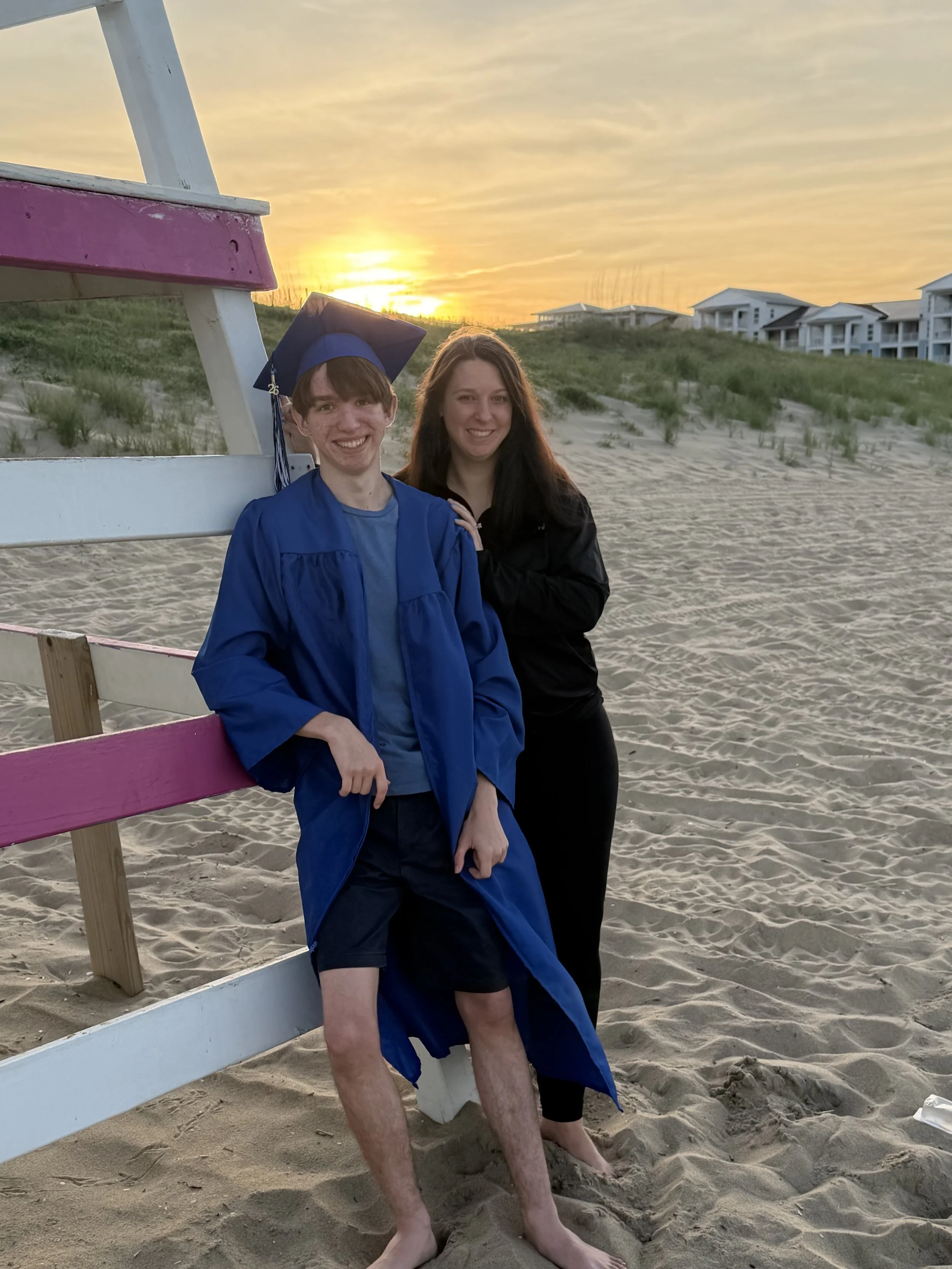 A young man in a Landstown High School graduation cap and gown standing on a sandy beach, smiling, with his parent next to him. The setting sun is in the background, casting a warm glow over the Virginia Beach in Sandbridge.
