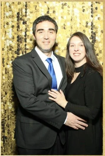 A man and woman standing together in front of a gold sequin backdrop, dressed in formal attire, smiling at the camera.