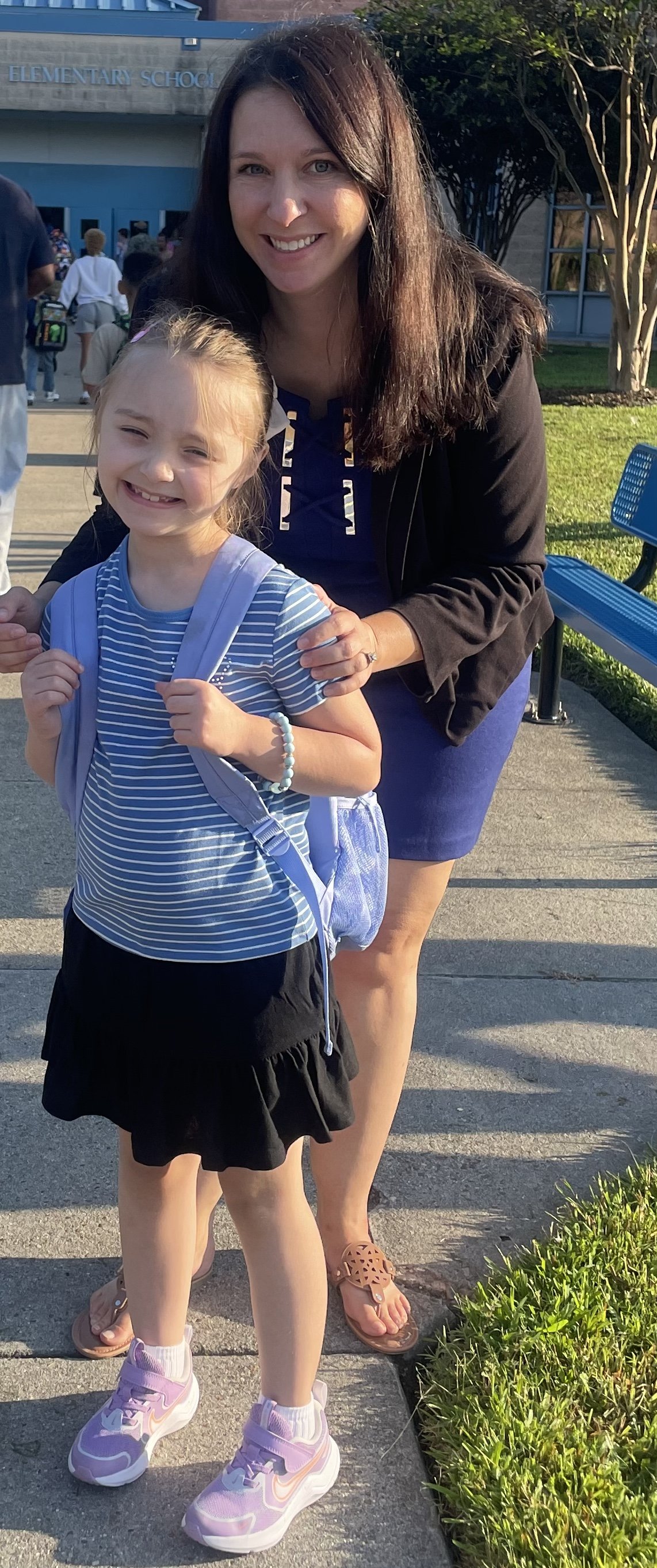 A woman and a young girl smiling outside in front of an elementary school with other children in the background. The woman has long dark hair and is wearing a black jacket and blue skirt, while the girl is dressed in a striped shirt, black skirt, and pink sneakers, and has a backpack on.