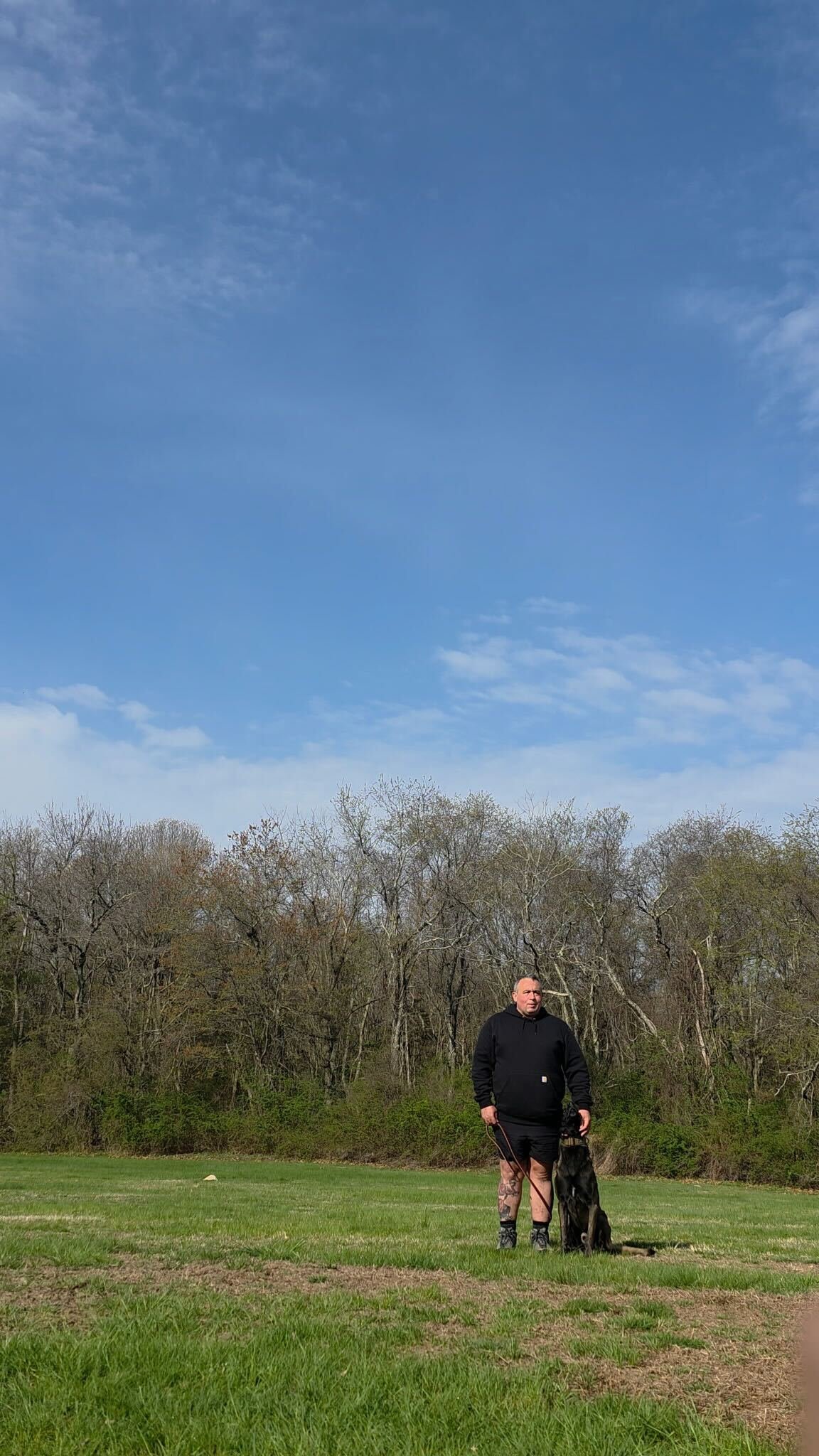 A man stands with a large black dog on a grassy field, with leafless trees in the background and a partly cloudy blue sky above.