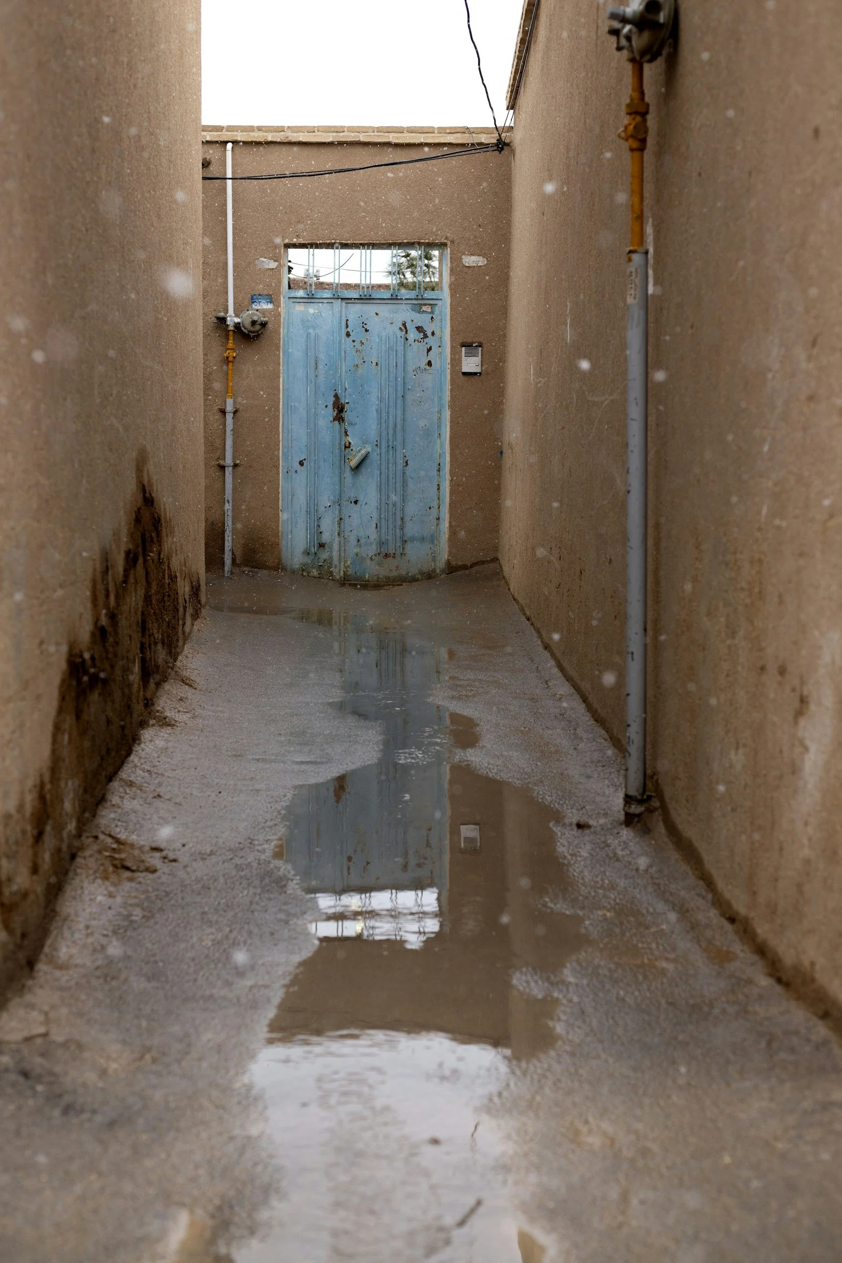 Narrow alleyway with muddy, wet ground and a puddle reflecting a metal blue door at the end, surrounded by beige walls and some exposed pipes.
