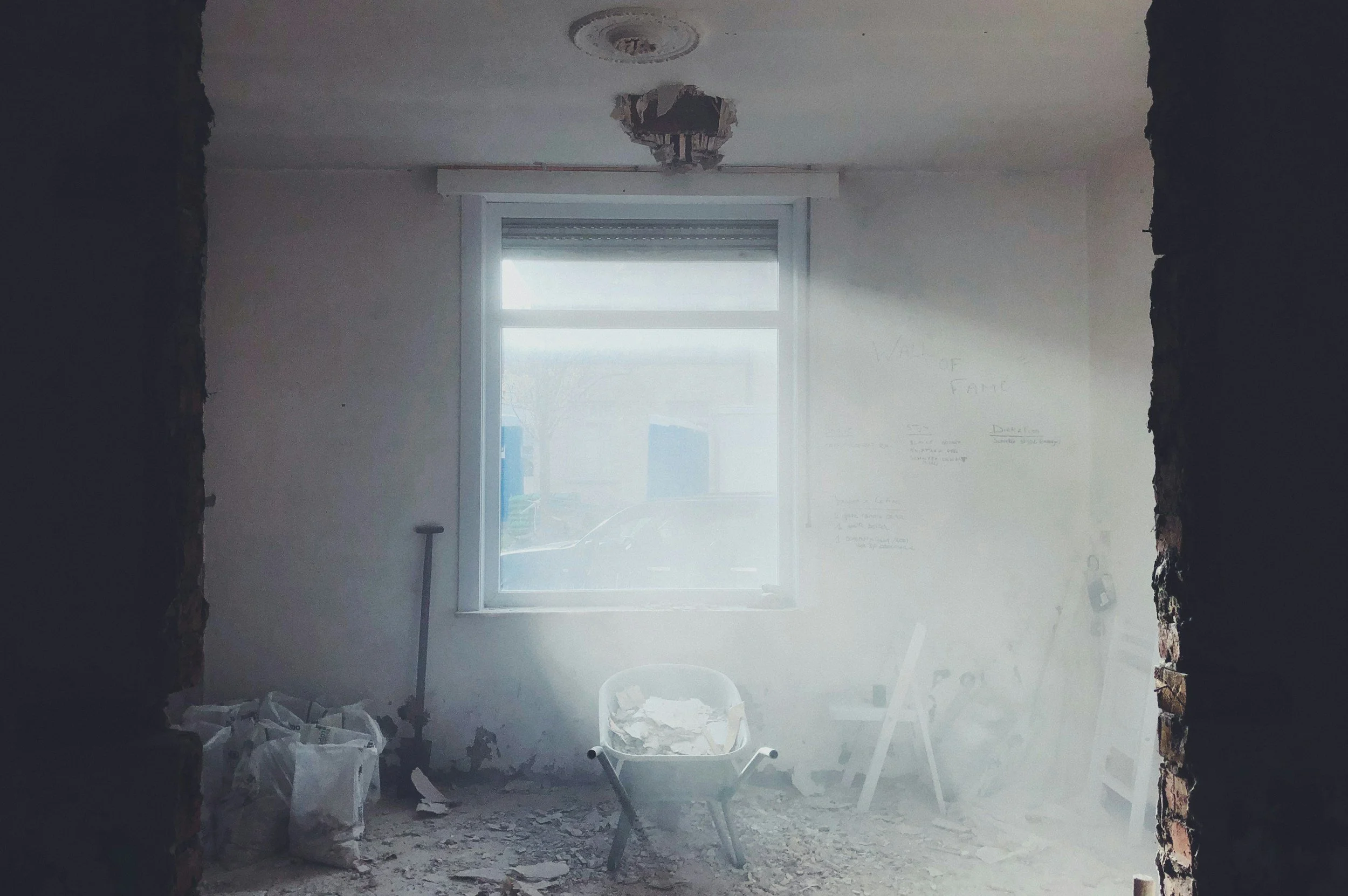 Room under construction or renovation with debris on the floor, an empty wheelbarrow, and a window letting in light, with exposed brick on the sides.