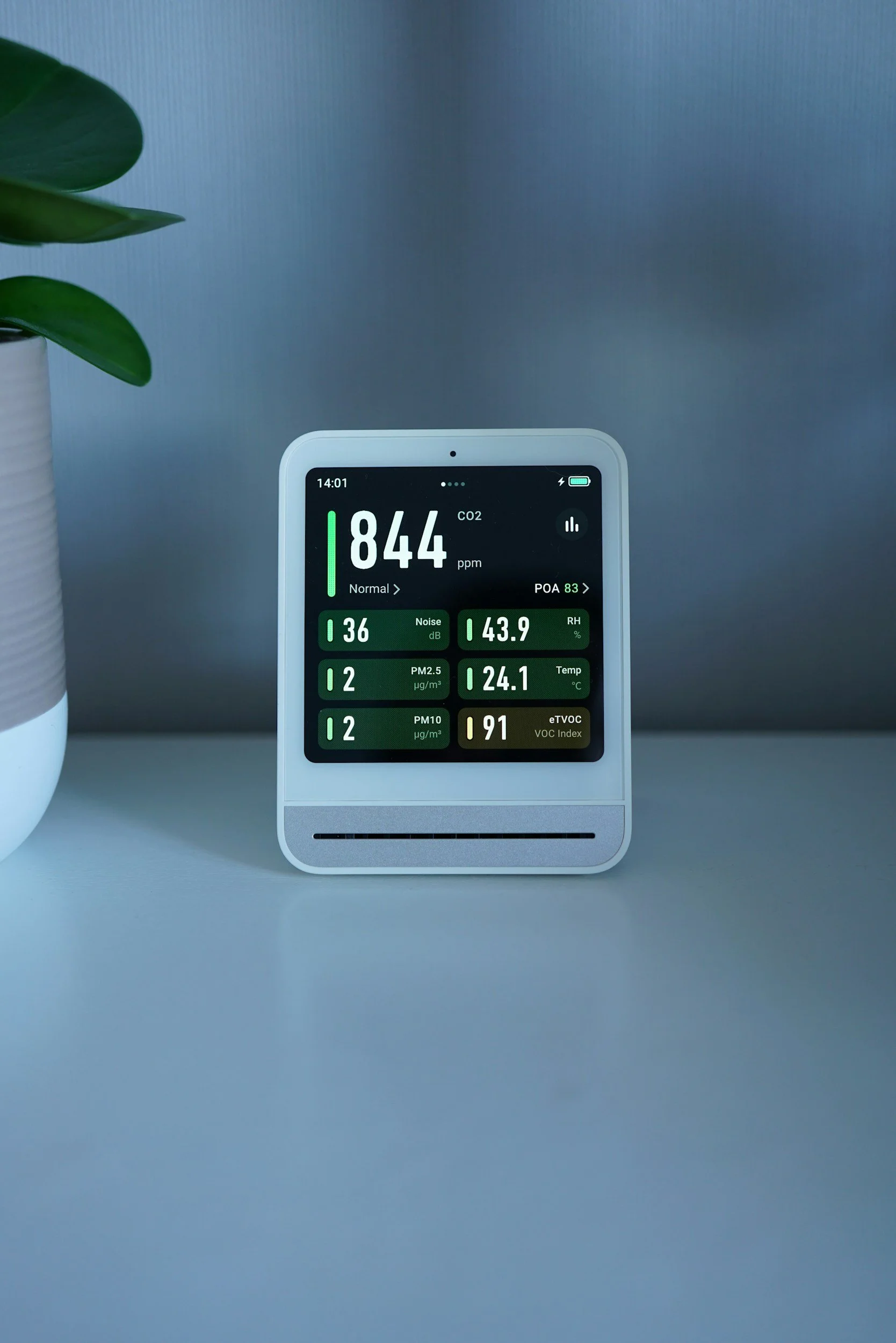 Air quality monitor displaying various environmental measurements such as CO2 level at 844 ppm, noise level at 36 dB, humidity at 43.9%, temperature at 24.1°C, and VOC index at 91, placed on a white surface next to a potted plant.