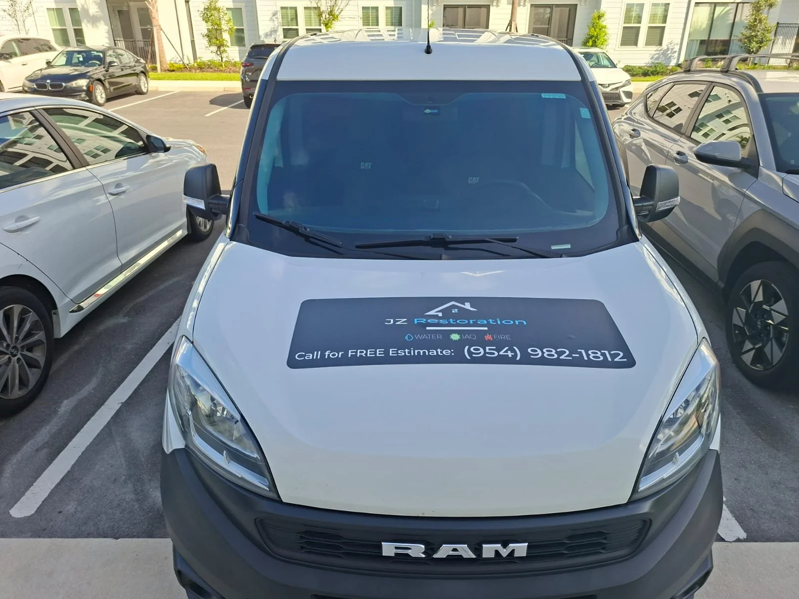 White RAM commercial vehicle with a black and blue advertisement on the hood for JZ Restoration, a water, IAQ, and fire restoration company, including a phone number for free estimates, parked in a lot surrounded by other vehicles and apartment buildings.