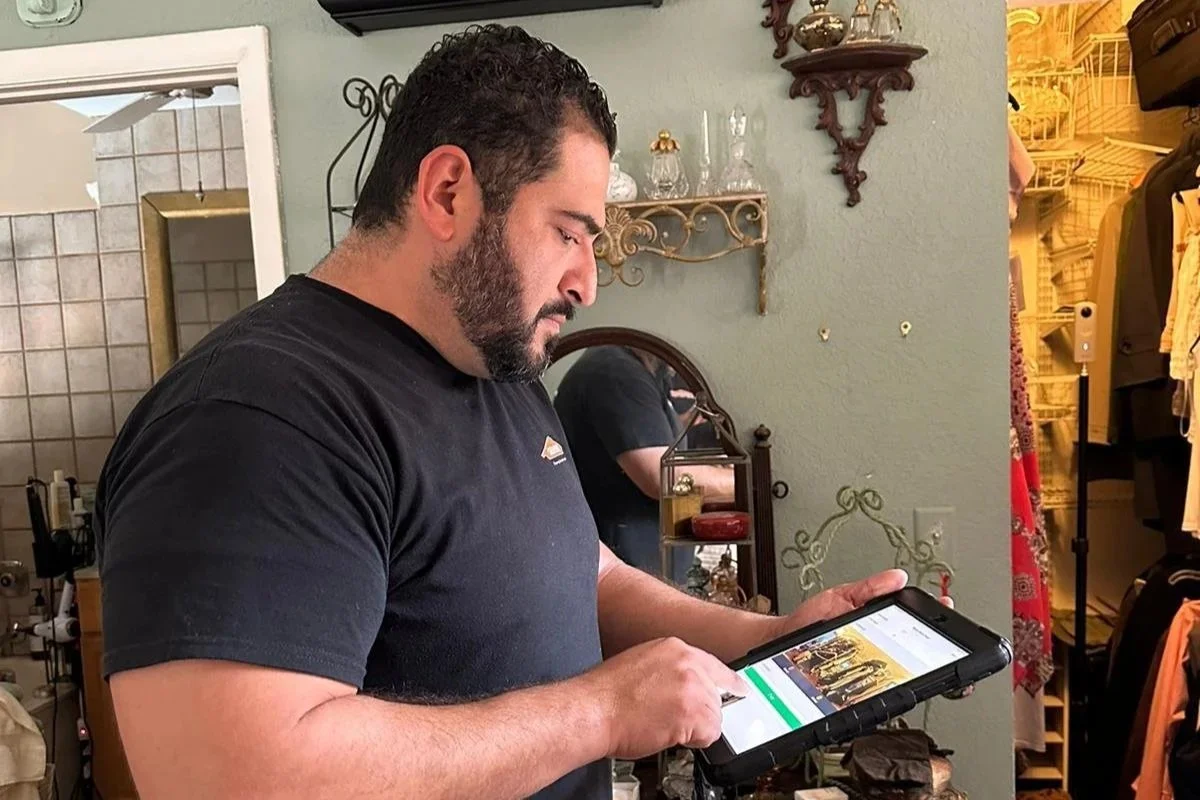 A man with dark hair and a beard, wearing a black t-shirt, standing in a room with vintage decor, looking at a tablet.