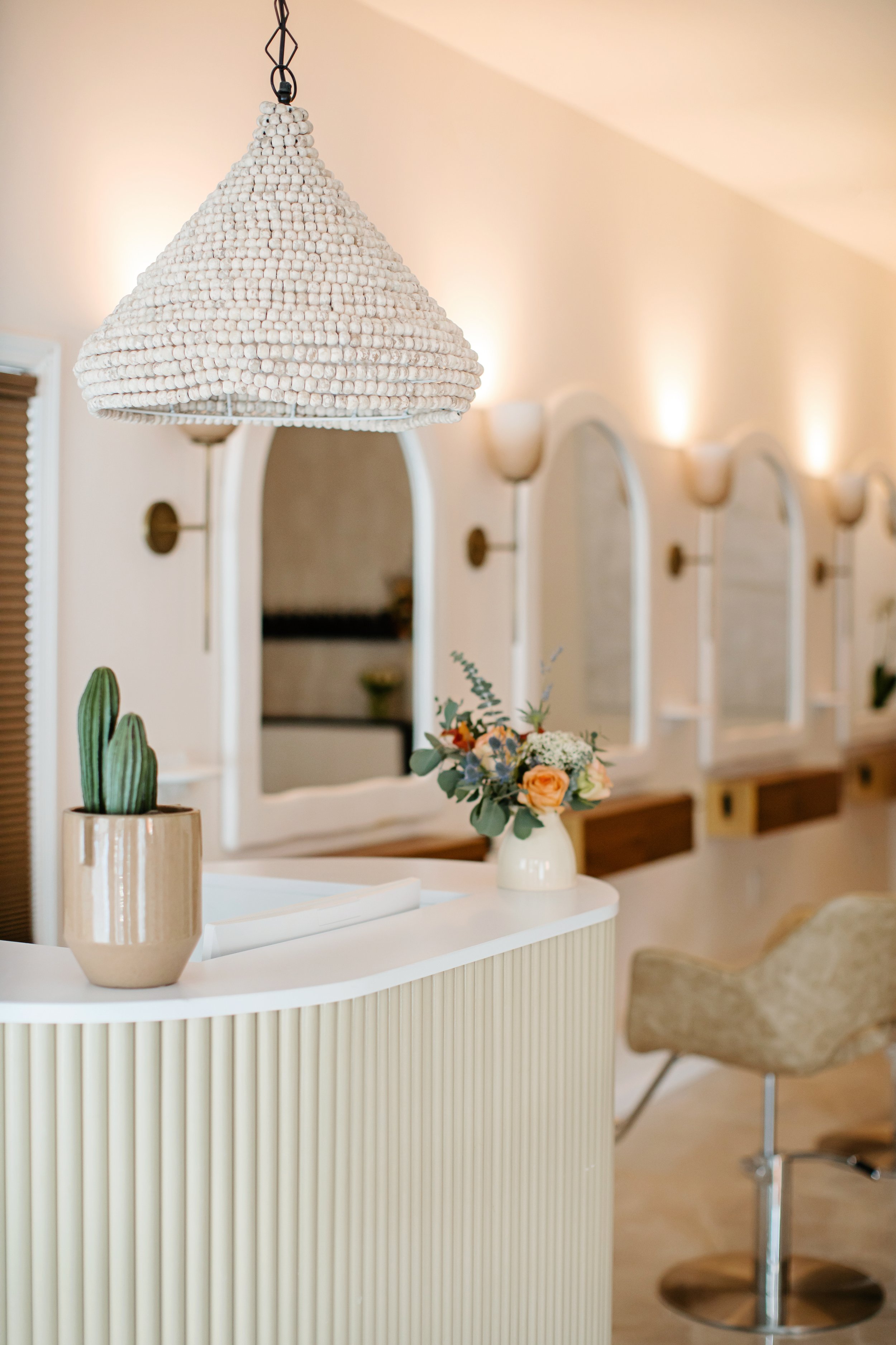 A white flowered lampshade hanging above a white curved desk with a beige vase holding green succulent plants, in front of a mirror and wall sconces in a brightly lit room with cream-colored walls.