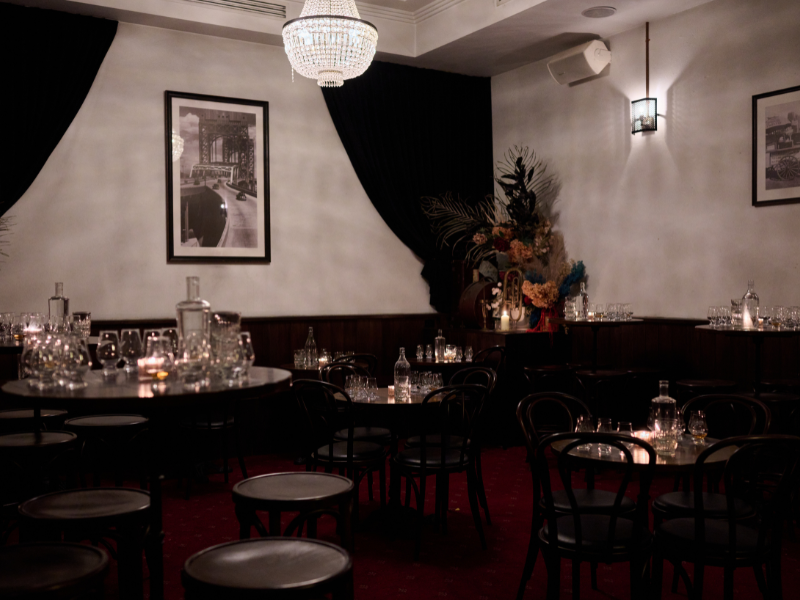 Elegant restaurant or event space with round tables, black chairs, glassware, bottles, and floral decorations, dim lighting, and framed black-and-white photographs on the walls.