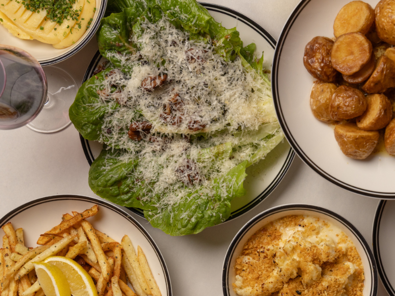 A dinner table with a plate of Caesar salad with parmesan cheese, a bowl of roasted potatoes, a bowl of French fries with lemon wedges, a bowl of macaroni and cheese, and a glass of red wine.