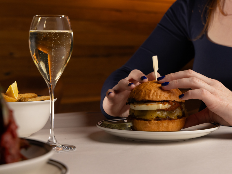 Person with navy long-sleeve shirt eating a large burger with pickles, onions, and cheese at a restaurant table, with a glass of sparkling beverage and a bowl of fried food nearby.