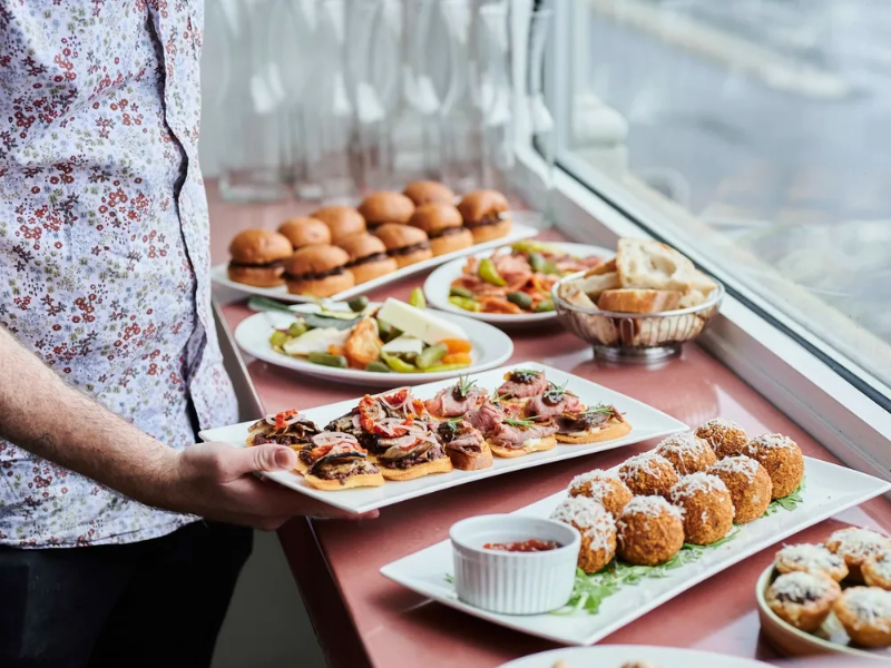 Person holding a plate of assorted canapés or appetizers near a window with a variety of other food items on a table, including bread, sliders, and fried snacks with dipping sauce.